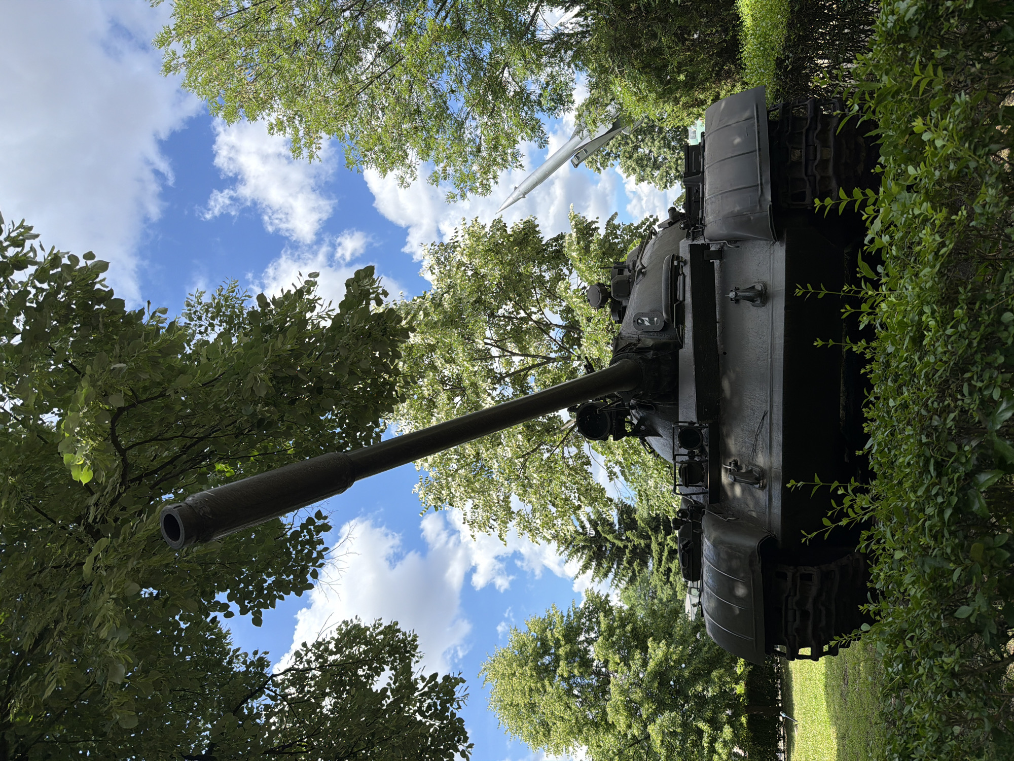 This is a full, eye-level shot of a dark, military tank parked on a green lawn, in front of a dense, green bush. The tank's barrel points diagonally upwards to the left, under the canopy of a large tree with green leaves. Behind the tank, a silver rocket is mounted on a stand, pointing upwards and to the right. The background shows a clear blue sky with scattered white clouds, and more green trees. The tank is identified as a T-55 tank and the rocket as a Scud missile. The scene is located at the Alley of the Soviet Army in Sofia, Bulgaria. There are no people visible in the frame.