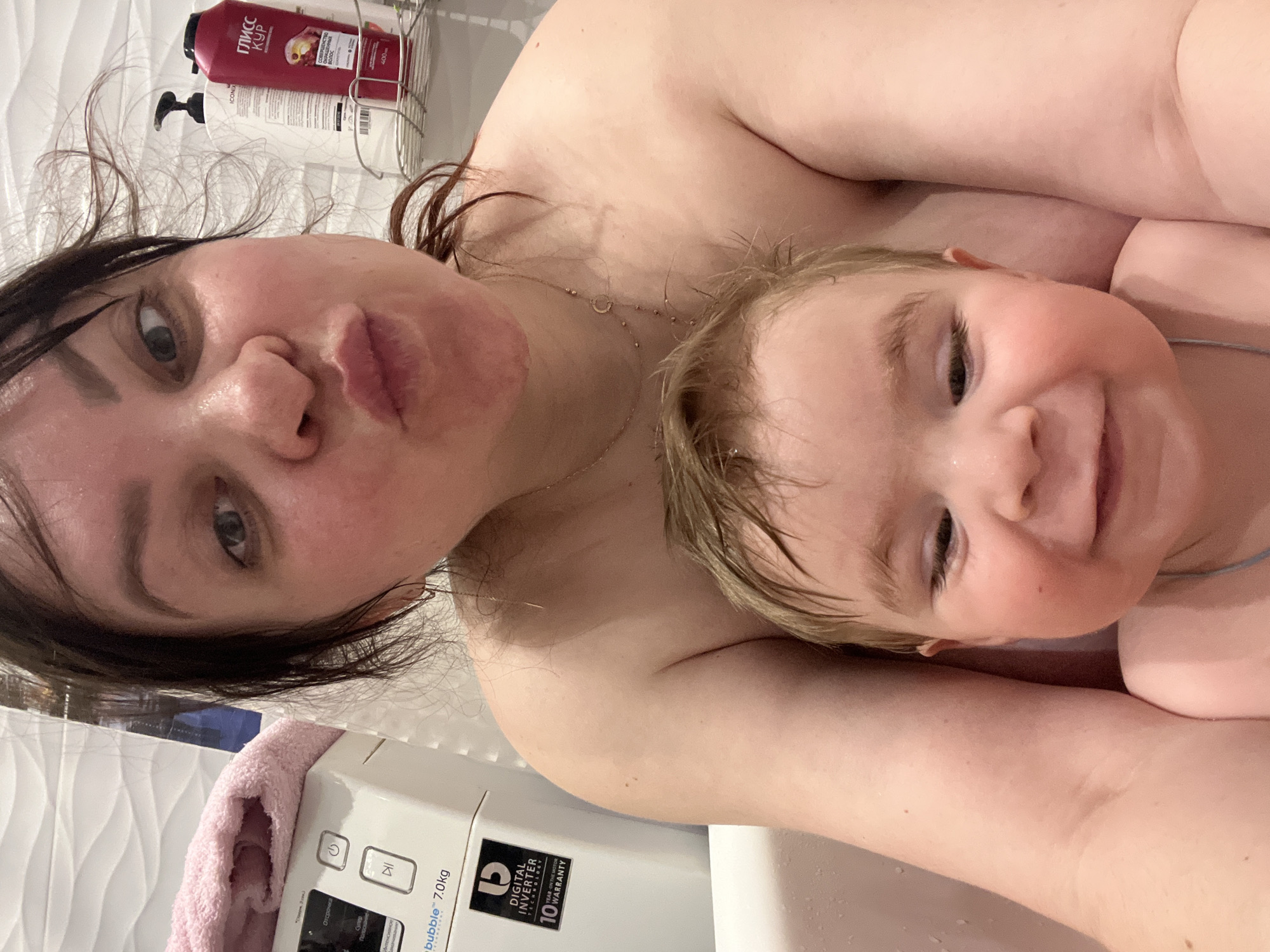 This image captures a tender moment between a mother and her young child in a bathroom setting, likely in Perm, Russia. The photo appears to be a selfie taken from a top-down perspective, possibly while they are in a bathtub or shower.
The woman, with wet, dark hair and a playful expression featuring pursed lips, looks directly at the camera. She wears a delicate gold necklace with small beads and a crescent moon pendant. Resting beside her, the child, with wet, light-colored hair, beams a wide, happy smile towards the camera. Both subjects have wet skin, suggesting they have just bathed or are still in the water.
In the background, behind the woman's head, a metal shower caddy holds two bottles: a red bottle labeled "ГЛИСС КУР" (GLISS KUR) with text "СОВЕРШЕНСТВО" (Perfection) and "400" (likely ml), and a white bottle with a black pump dispenser and the brand name "ICONEX". Below the subjects, a white washing machine is visible, displaying buttons and text including "Отсрочка" (Delay/Postpone), "bubble™ 7.0kg", "DIGITAL INVERTER", and "10 WARRANTY YEAR ON THE MOTOR". A soft pink towel is draped over the washing machine. The wall behind them features white, subtly textured tiles. The overall mood of the image is intimate, joyful, and relaxed.