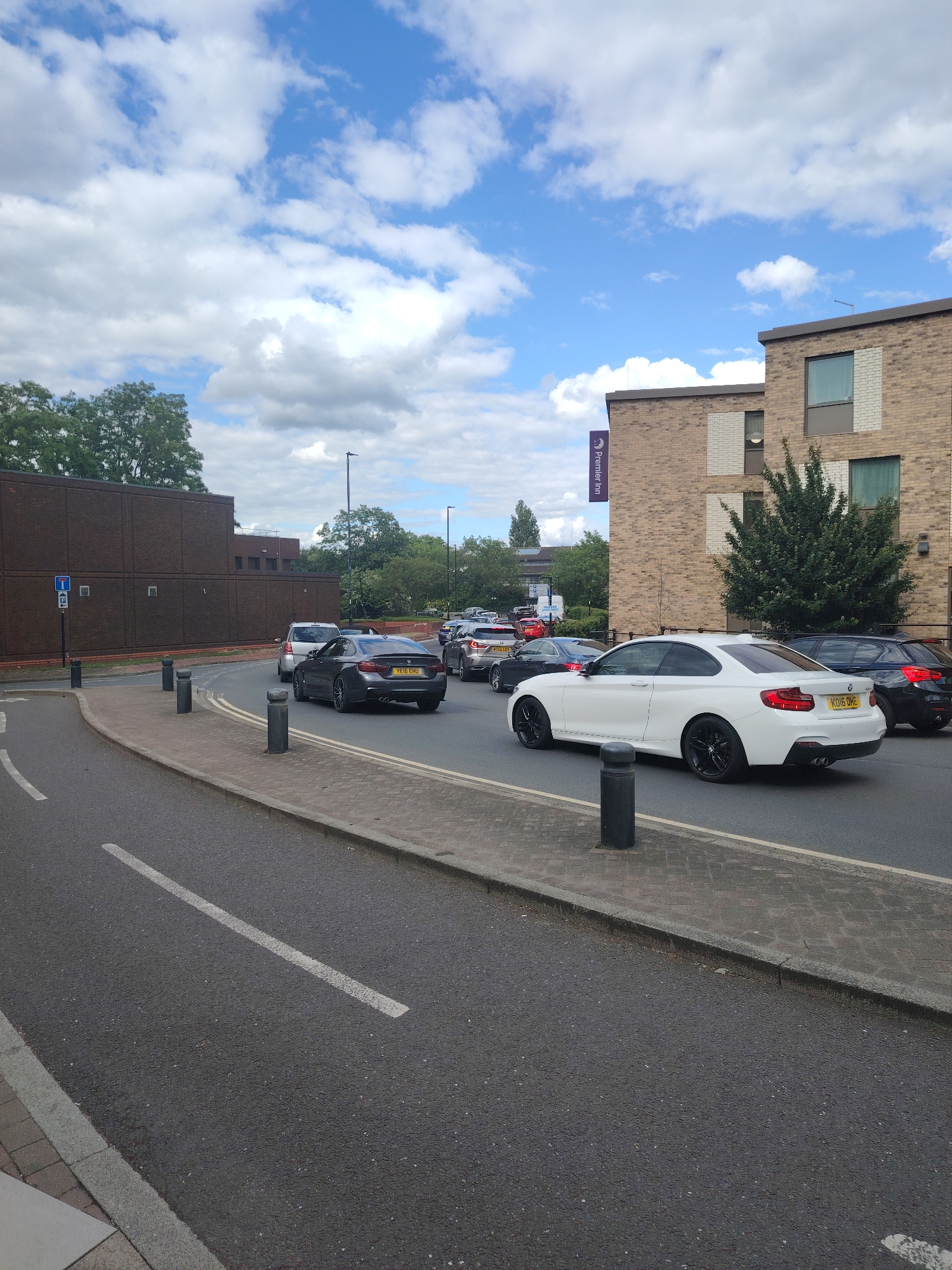 This image depicts a daytime scene on Fleming Way in Peterborough, United Kingdom, looking northeast towards the Peterborough City Centre area (PE1 1AP). A multi-lane asphalt road curves from the left foreground towards the right midground, occupied by a queue of vehicles.
In the foreground, a white BMW 2 Series Coupe (registration KO16 OHE) is positioned on the right side of the road, facing away from the camera. Behind it, further back in the traffic queue, a dark grey BMW (registration YE16 EHU) is also visible. Other vehicles are intermittently visible in the queue further down the road.
To the immediate right of the road, a paved pedestrian walkway is present, separated from the roadway by a series of dark grey bollards. This walkway runs alongside a multi-story brick building, identified as the Premier Inn Peterborough City Centre by a prominent purple vertical banner sign displaying "Premier Inn". The building features various windows and some lighter brick accents.
To the left of the road, a large, rectangular, dark brown brick building stands. This structure is consistent with the Peterborough Magistrates' Court complex. Adjacent to this building and the road, a blue road sign displays white symbols for a taxi rank (a capital 'T') and parking (a capital 'P').
The background includes several deciduous trees with green foliage. The sky above is partially cloudy, featuring scattered white cumulus clouds against a blue backdrop. No discernible people are individually identifiable within the frame.