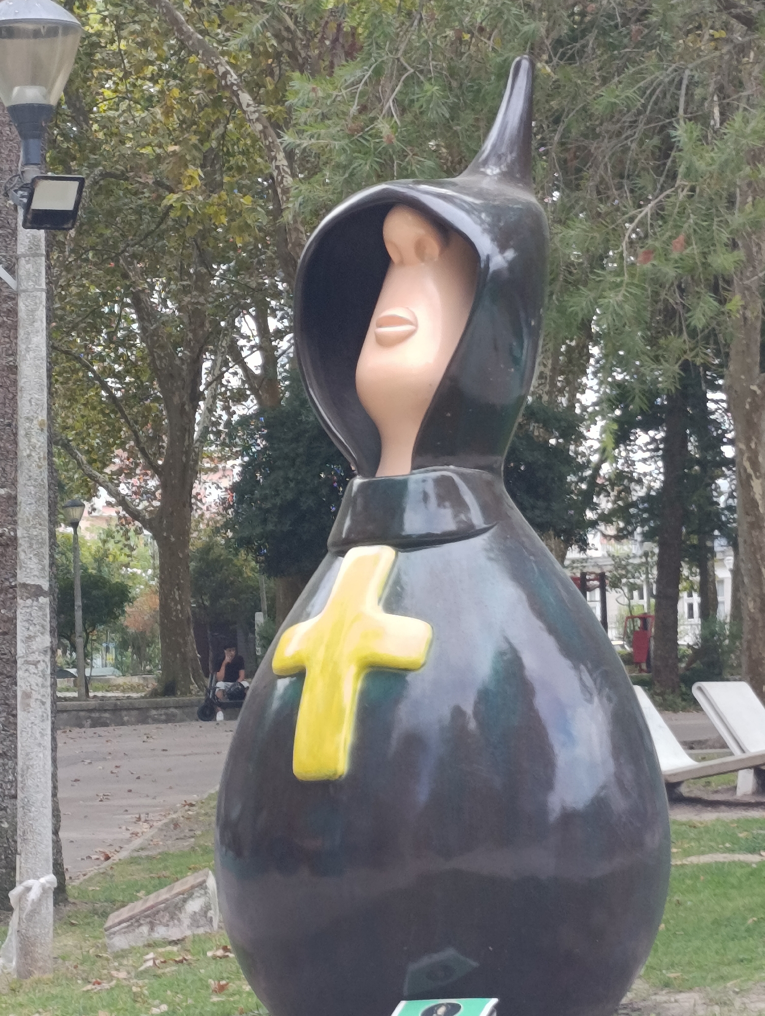 This image shows a large, dark, egg-shaped statue with a pointed hood and a stylized, pale face with exaggerated features. A bright yellow cross is attached to the front of the statue. The statue is situated in an outdoor public space, likely a park, with trees and a paved walkway visible in the background. There is a lamppost on the left side of the image. In the background, a person is sitting on a bench. This statue is part of the "Alegro Setúbal" shopping center's outdoor area, located in Setúbal, Portugal.