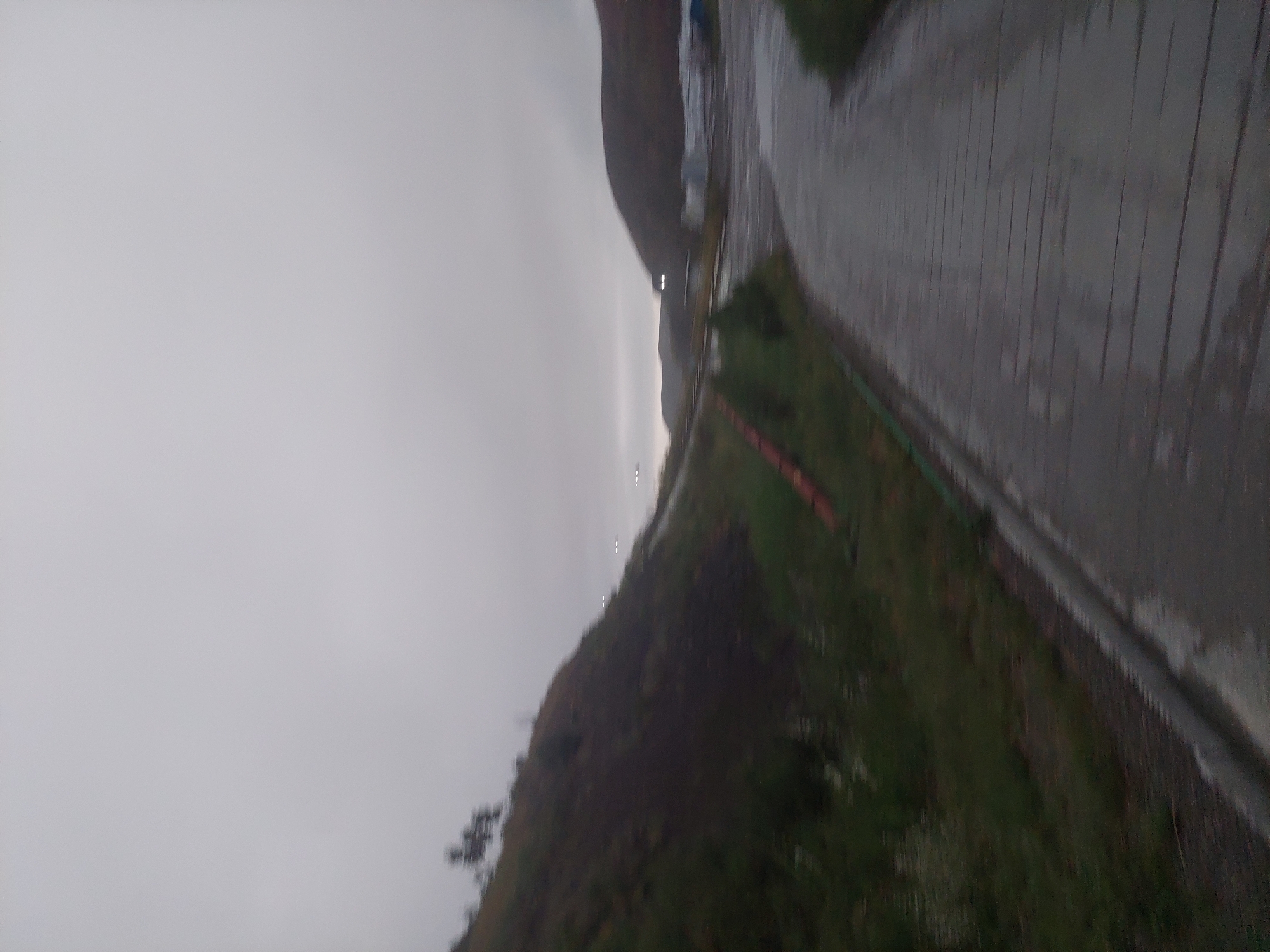 The image depicts a gray, overcast sky above a landscape that includes a steep, grassy hill on the left and a paved road curving towards the right. Sparse vegetation grows on the hillside, and what appear to be distant lights are visible on the horizon. Structures, possibly industrial buildings, are partially visible in the middle ground. The road appears wet, suggesting recent rain. The scene has a muted, somber atmosphere. The location is Bordunskiy, Kyrgyzstan.