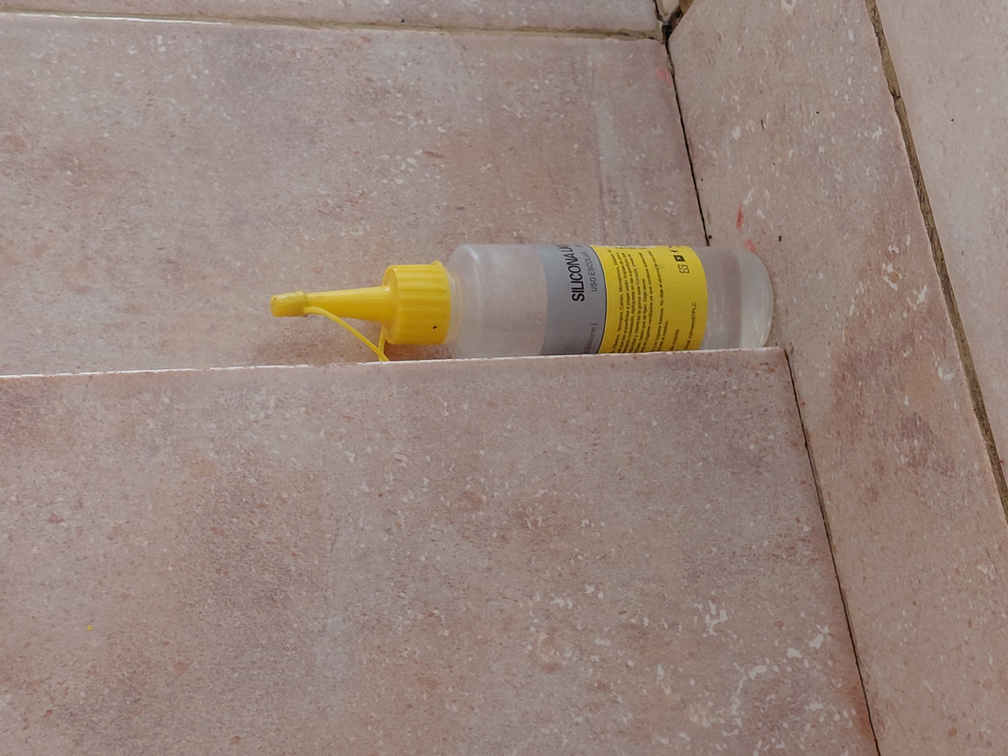 The image, captured in Santa Ana, Venezuela, features a clear plastic squeeze bottle with a vibrant yellow cap and nozzle, lying on a tiled surface. The bottle contains a translucent liquid, possibly glue or silicone. It is positioned horizontally across a grout line, resting on light brown or beige tiles with a speckled, mottled texture. The tiles appear to form a corner or a step, suggesting an indoor setting such as a floor or staircase.
On the side of the bottle, a grey label with black text is visible. Key text includes "SILICONA LÍQUIDA" (Liquid Silicone) and "USO ESCOLAR" (School Use), indicating the product's nature and intended application. Below this, a yellow sticker contains smaller, mostly illegible black text, which appears to be instructions or warnings in Spanish, with partial words like "adherirse" (to adhere), "superficies" (surfaces), "secar" (to dry), "fijas" (fixed), and "ventilados" (ventilated) being faintly discernible. A small barcode-like symbol and the text "ES 1" are also visible on this yellow sticker. The bottle itself has a string attached to the nozzle, possibly for a cap or to prevent loss. The overall scene is static, with no human activity depicted.