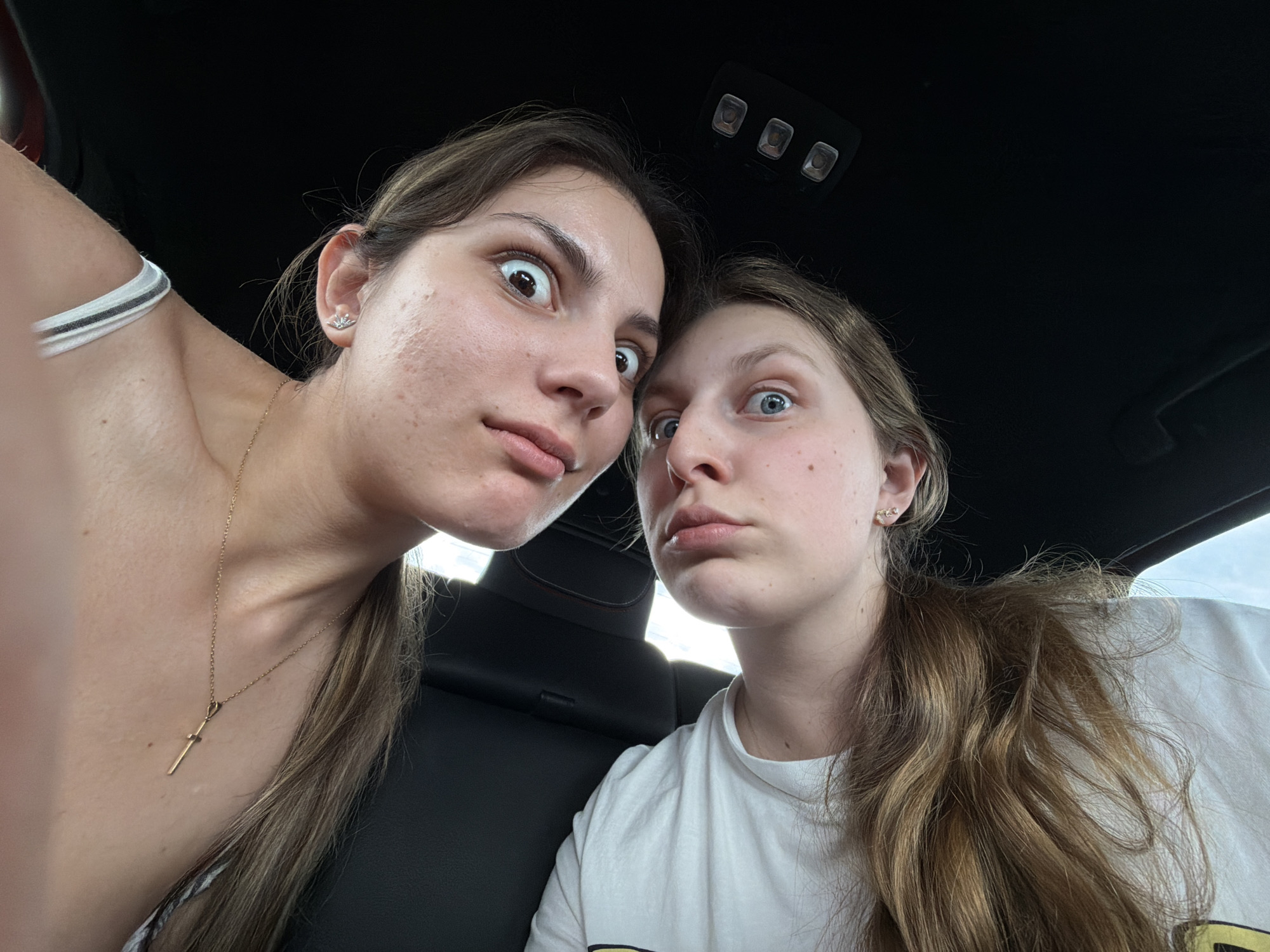 Two young women are inside a car. Both are making exaggerated facial expressions with wide eyes and pursed lips. The woman on the left has dark hair, a gold necklace, and is wearing a white top with a white strap visible. The woman on the right has light brown hair and is wearing a white t-shirt. The car's interior includes black seats and overhead lights. The location is unidentifiable as Chokoba, Bulgaria from the given image.