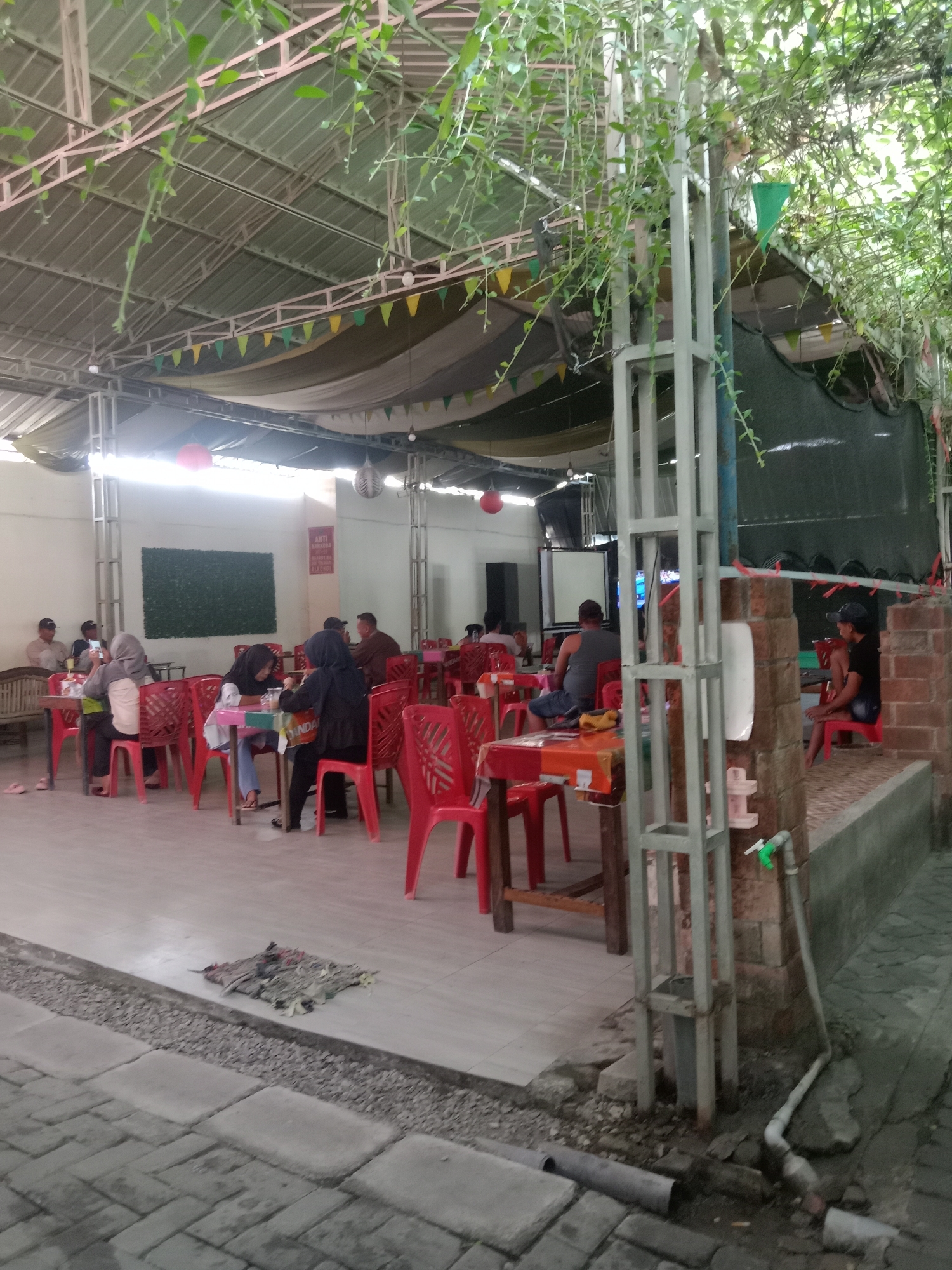 The image depicts an outdoor dining area, likely a restaurant or cafe, with several tables and red plastic chairs occupied by patrons. The space is covered by a metal roof supported by a steel frame, with hanging plants adding a natural element. Colorful pennant banners are strung across the ceiling, suggesting a festive or casual atmosphere. In the background, a large green wall covered in artificial foliage is visible, along with a television screen. A water faucet and plumbing are situated near a brick wall on the right side of the frame. The ground is paved with tiles and gravel. The scene is set in Kemamang, Indonesia, a location implied by the provided information.