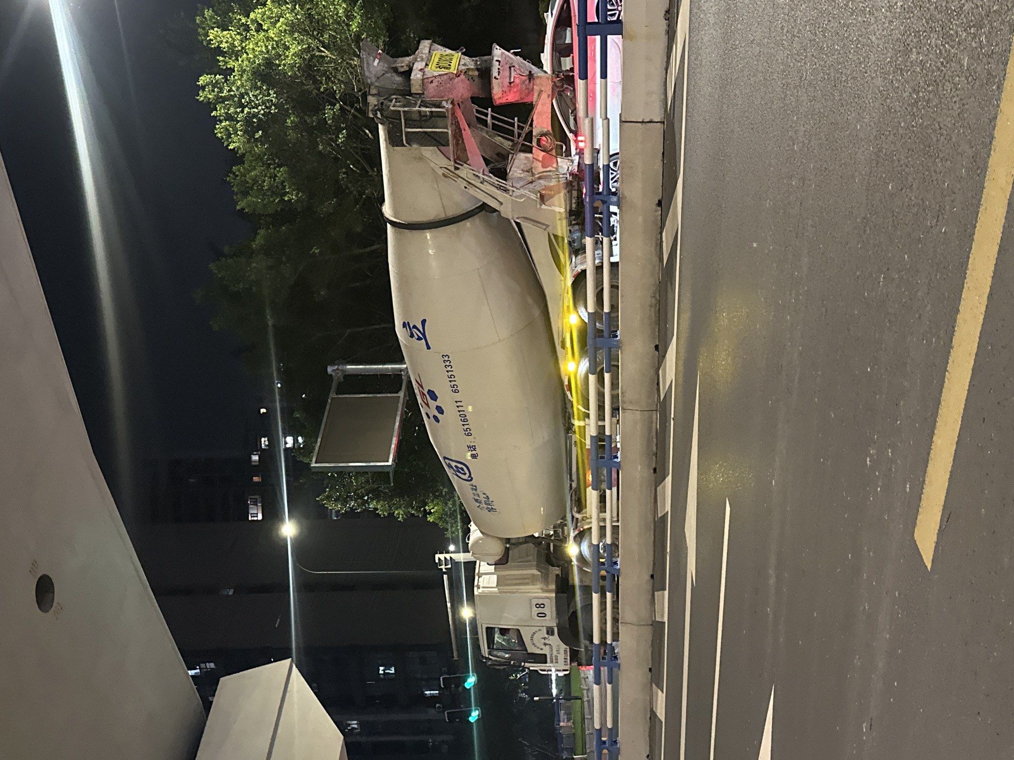 A concrete mixer truck is captured at night on a road in Chongqing High-tech Zone, China. The truck, a white cab with a white drum, is positioned on the right side of the image, its drum slightly tilted. The truck's side displays Chinese characters and a phone number. The driver is not visible in the cab. The truck is traveling on a multi-lane road with a concrete barrier separating the lanes and a pedestrian walkway. The road surface is wet and reflects the streetlights and vehicle lights. Yellow and white road markings are visible. In the background, trees are illuminated by lights, and a tall building with lit windows is present. A traffic light with green signals is visible to the left of the truck. The overall scene depicts an urban environment at night, with a focus on the movement of a construction vehicle.