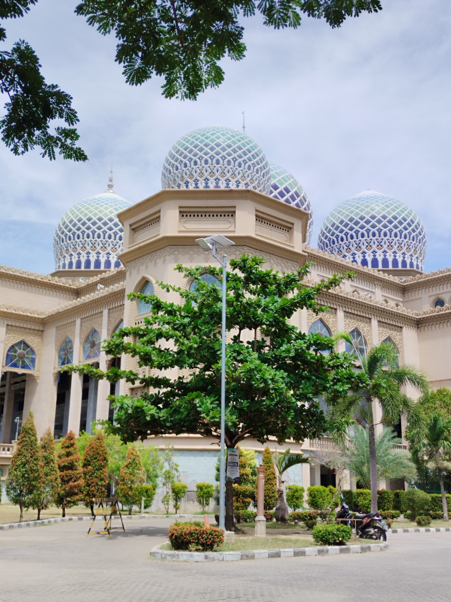 A grand mosque with ornate Islamic architecture is the central subject of this brightly lit daytime image, set in Lhokseumawe, Indonesia. Multiple large domes, predominantly blue, white, and green with intricate geometric patterns, including a diamond-like lattice, crown the beige-colored structure. The mosque features arched windows, decorative relief work on its walls, and visible columns.
In the foreground, a lush, leafy green tree partially obstructs the view of the mosque, while a tall, grey lamppost, which appears to be solar-powered, stands prominently. The ground consists of a paved area, likely a courtyard or parking lot, bordered by kerbs and interspersed with well-maintained landscaping. Various types of greenery are present, including shorter bushes, slender coniferous trees, and palm trees, creating a verdant environment around the building.
To the right of the frame, a motorcycle is parked near the kerb. Further back, a black and yellow barrier can be seen. The sky above is mostly clear with a few wispy clouds, suggesting bright, sunny weather. A small, white sign is attached to the lamppost, with the Indonesian word "HIMBAUAN" (meaning "appeal" or "advice") partially legible. The scene is calm and devoid of people, emphasizing the architectural details and the natural surroundings.