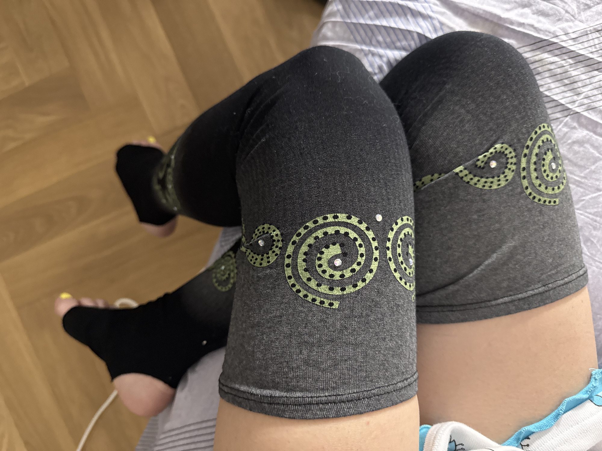 A first-person, medium shot depicts a person's legs from the upper thighs down to the feet. The person is wearing dark gray knee-high leggings with green circular designs that include small, sparkling embellishments. The feet are clad in black, footless tights or socks. The person is sitting on a bed with a striped white and gray sheet. The background includes a wooden floor. The location, Moscow, Russia, is not discernible from the image.