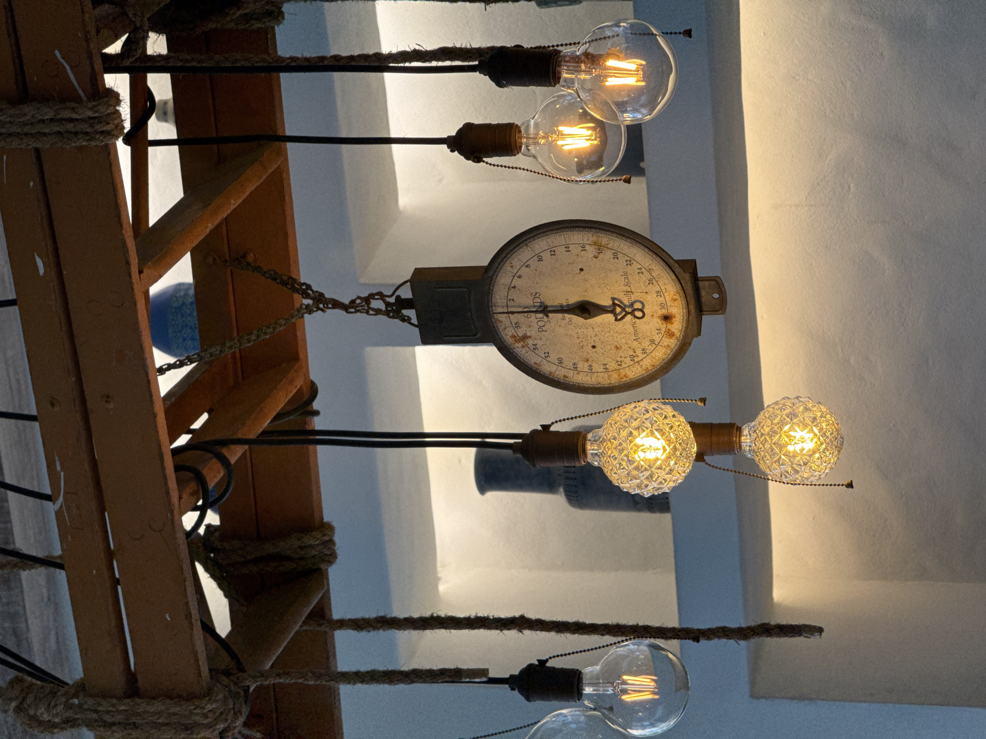The image shows a close-up of an industrial-style hanging light fixture and a vintage scale in what appears to be a restaurant or cafe setting in Hua Hin City Municipality, Thailand. The fixture is made of rough-hewn wood beams from which several incandescent light bulbs are suspended by rope and chain. One of the bulbs has a faceted glass shade, while others are clear Edison-style bulbs. The vintage scale, which hangs from a chain, has a circular face with markings indicating pounds and a central pointer. The background shows a white wall with recessed shelving, where two dark-colored vases are visible. The lighting is warm and ambient, emanating from the bulbs. There are no people present in the image.
