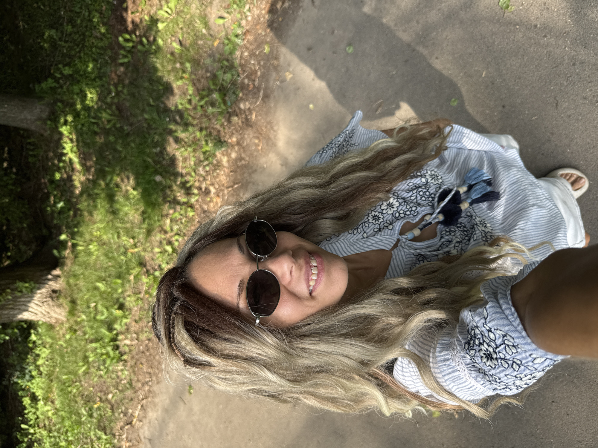 A woman is taking a selfie while walking on a path in a park. She has long, wavy blonde and brown hair and is wearing sunglasses, a blue and white striped shirt with embroidered details and tassels, and white shorts. The path is made of asphalt, and there is grass and trees in the background. The sun is casting shadows on the ground. The location is in the city of Rusanovo, Russia.