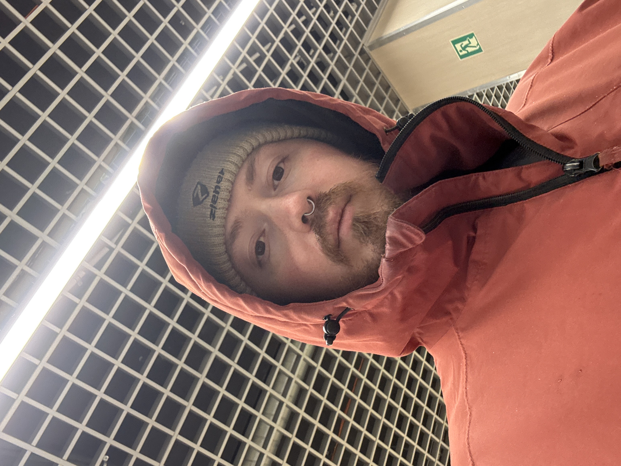 A person wearing a beanie and a red hooded jacket is looking at the camera. The person has facial hair and a nose ring. The background consists of a grid-like ceiling with a bright light fixture. An emergency exit sign can be partially seen in the upper right corner. The scene may be in a public space. There is no information to indicate that the picture was taken in Chaikovsky, Russia.