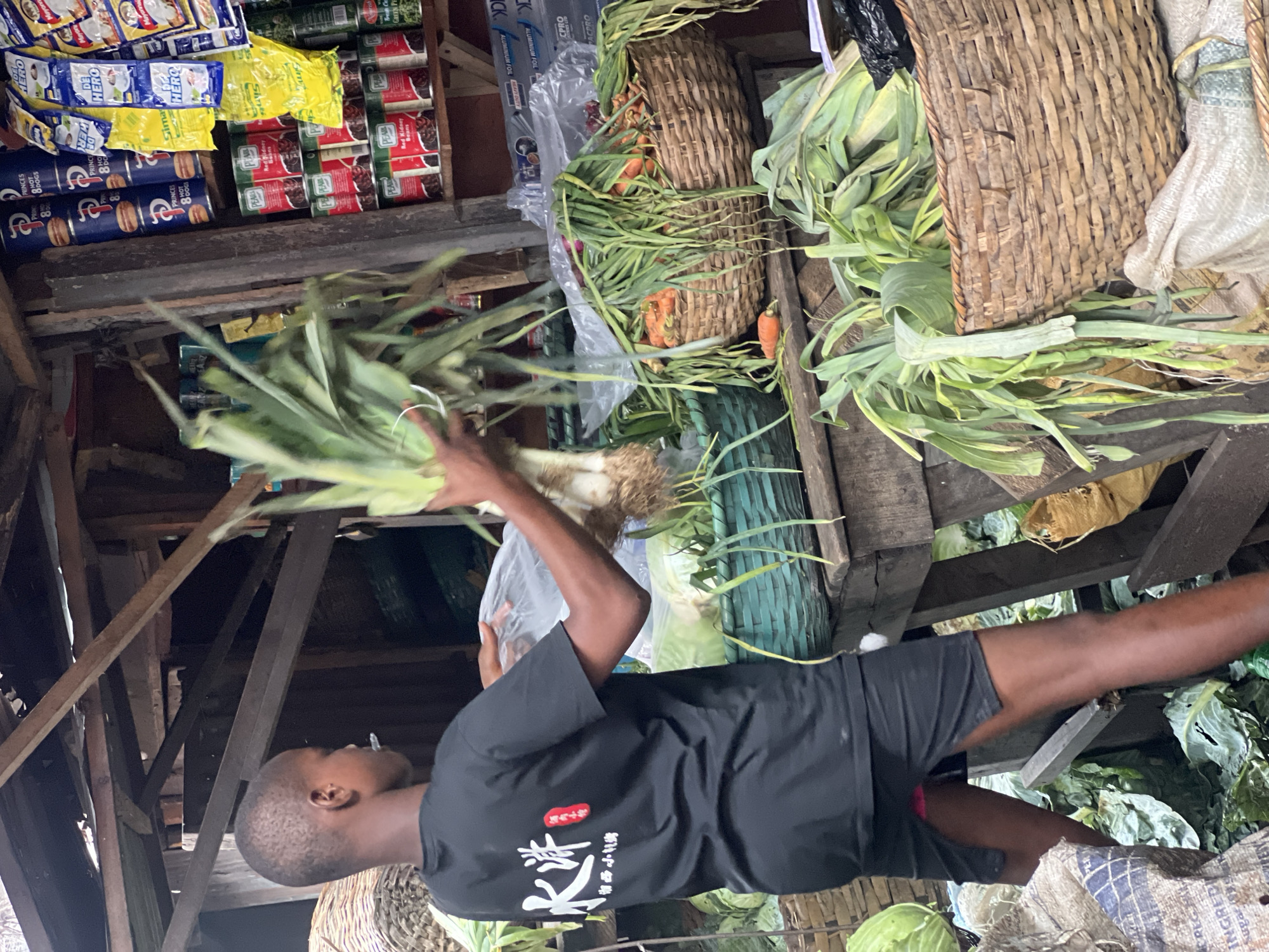 A street vendor in Nkpor, Nigeria, is seen holding a bundle of green leeks. The vendor, a young man with dark skin, is wearing a black t-shirt with white and red Asian script and black shorts. The vendor appears to be restocking his stand with the leeks. He is standing near a wooden structure that serves as a display for canned goods and packages of food products. Baskets of produce, including carrots, are visible behind the vendor. Cabbages and bags are on the ground.
