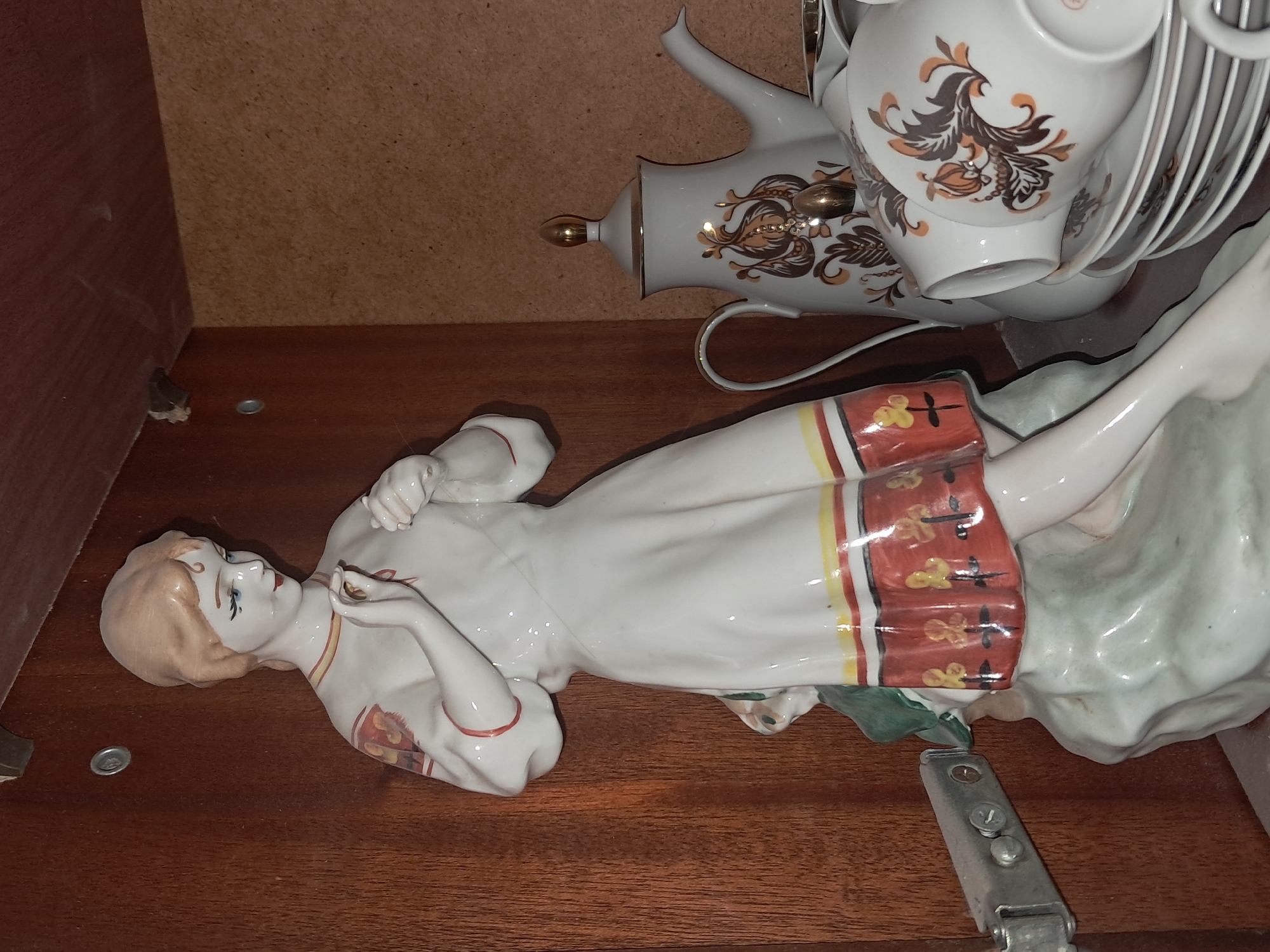 This media file showcases a collection of porcelain decorative items, primarily a tea set and a figurine, arranged on a wooden shelf. The tea set, positioned in the upper right portion of the frame, includes a teapot adorned with gold and brown floral patterns, along with stacked cups and saucers. To the left of the tea set, a figurine of a woman lies horizontally, her body extended to the left. The figurine is depicted with a pale complexion and blonde hair, wearing a white dress with a red, yellow, and black striped skirt that has decorative gold motifs. Her arms are bent, with one hand near her face. The background consists of a light brown particleboard surface. The overall scene appears to be indoors, likely on a shelf within a cabinet or display unit. The lighting suggests an indoor environment, with no indication of natural light or specific time of day. The figurine's pose and the placement of the tea set suggest a decorative display rather than an active scene. No visible text is present in the image.