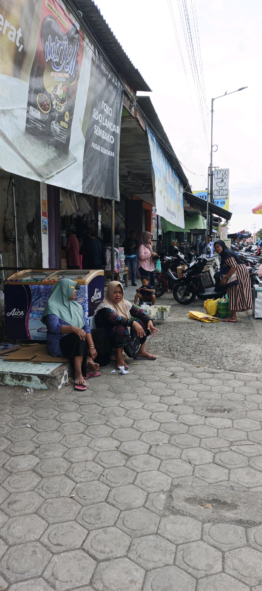 The image depicts a street scene in Kasiyan, Indonesia, likely a market area, characterized by stalls, motorbikes, and pedestrian activity. In the foreground, two women are seated on a raised concrete step. The woman on the left wears a light green hijab and a striped blue and white shirt with dark pants. The woman on the right wears a light brown hijab and a dark, patterned dress. Between them, a commercial freezer with "Aice" branding is visible.
The background reveals a row of shops with awnings extending over the sidewalk. Various signs are displayed, including one for "TOKO SIDO LANCAR SEMBAKO PASAR REBOAN" and another for "BAKSO MIE AYAM". Several motorbikes are parked along the street, and other individuals, including a child crouching, are present in the mid-ground. The ground is paved with hexagonal concrete blocks. Overhead, power lines are strung across the sky, with a street lamp visible on the right. The overall atmosphere suggests a busy, everyday commercial environment.