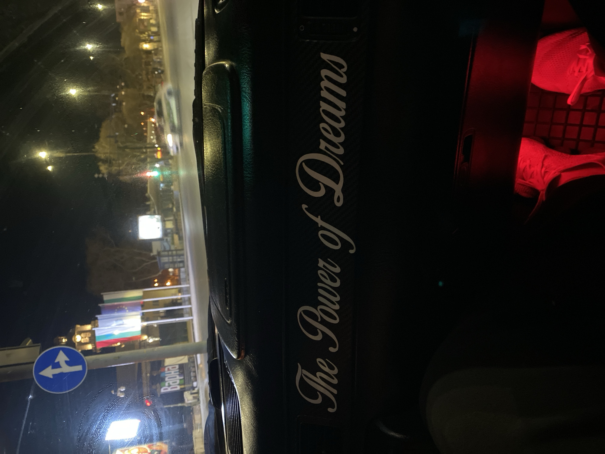 The image shows an interior view from what appears to be a vehicle in Varna, Bulgaria, at night. On the right, partially illuminated in red, is a seat or storage compartment with white fabrics or items within. The dashboard prominently displays the text "The Power of Dreams" in elegant lettering. Through the windshield, the exterior is visible. There is a blue road sign with a left-pointing arrow. Further in the background, Bulgarian and possibly EU flags are seen. Streetlights and illuminated buildings suggest an urban setting. The sign to the right says Varna in Bulgarian Cyrillic - Варна.