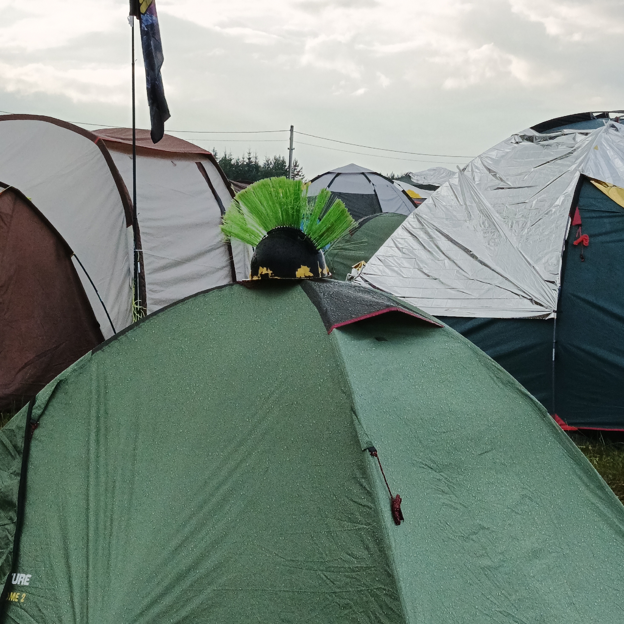 The image depicts a campsite with multiple tents set up outdoors. In the foreground, a green tent is prominent, with a black helmet adorned with bright green spikes resting on top of it. In the background, other tents are visible, including a brown and white tent, a grey and black dome tent, and tents covered in reflective material. A flag on a pole is also visible in the upper left portion of the image. The sky is overcast with clouds. The scene is likely a music festival or camping event, given the presence of a unique headwear item and the arrangement of tents. The location is stated to be Lagunovo, Russia.