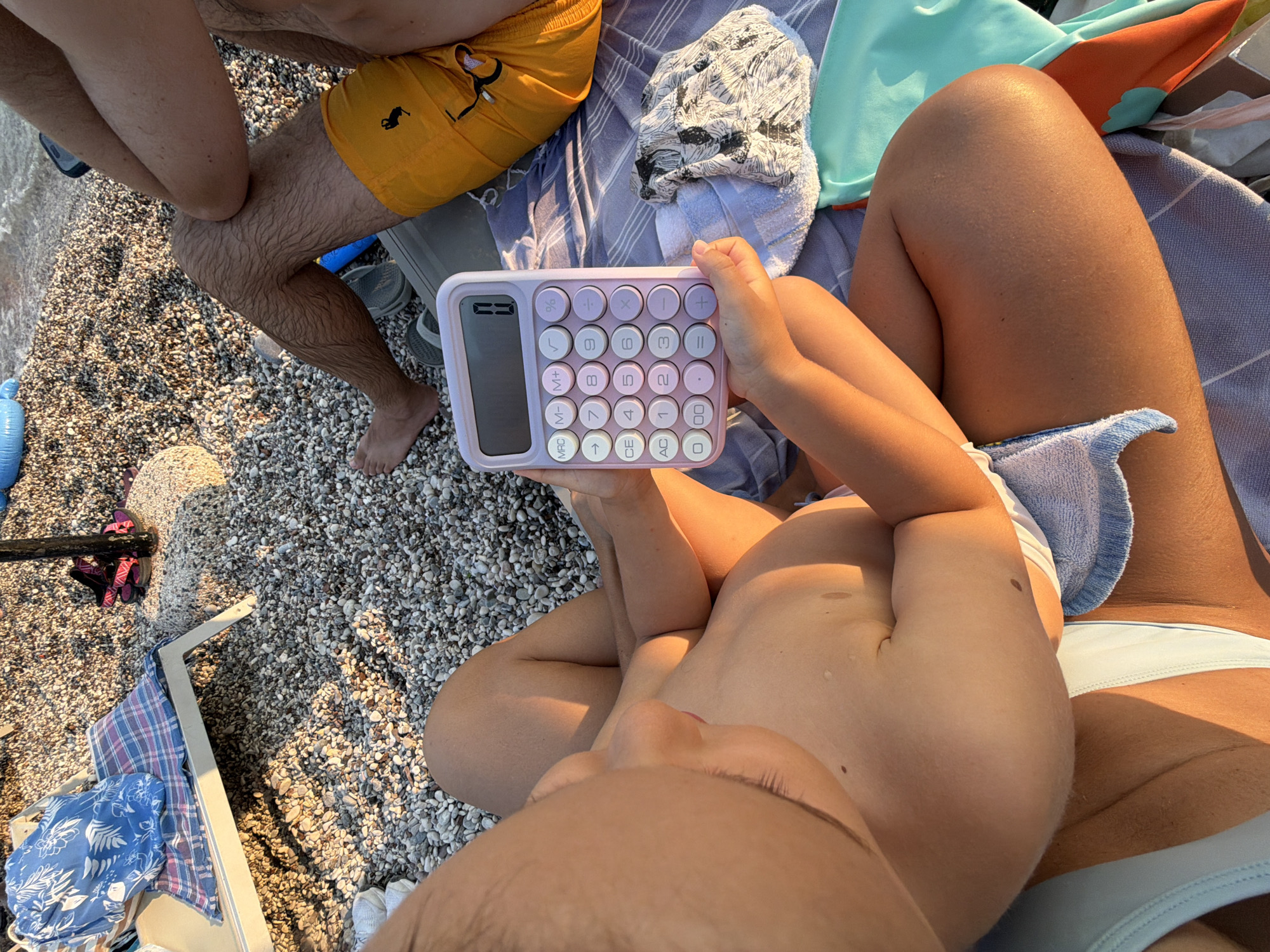 This is a high-angle, close-up shot of a group of people relaxing on a pebble beach in Budva, Montenegro. The sun is shining, casting long shadows, suggesting it is late afternoon or early morning.
In the foreground, a child is holding a pastel purple calculator with a digital display. The calculator has round buttons with numbers and symbols. The child's hands are visible, gripping the device. Part of the child's torso and legs are also in view, with smooth, tanned skin.
To the left of the child, an adult's bare legs are visible, resting on the pebbles. They are wearing yellow swim trunks with a black silhouette design. Behind them, more legs are visible, indicating at least one other person is present.
The subjects are sitting on a collection of towels and beach accessories. A striped towel is prominent, along with a patterned fabric and a bright teal-colored item, possibly a bag or clothing. The beach is covered in small, smooth pebbles, and the edge of the sea is visible to the far left.
The overall mood is relaxed and casual, typical of a beach vacation. The sunlight highlights the textures of the pebbles and the skin of the people. The scene captures a moment of leisure and play on a sunny day by the water.