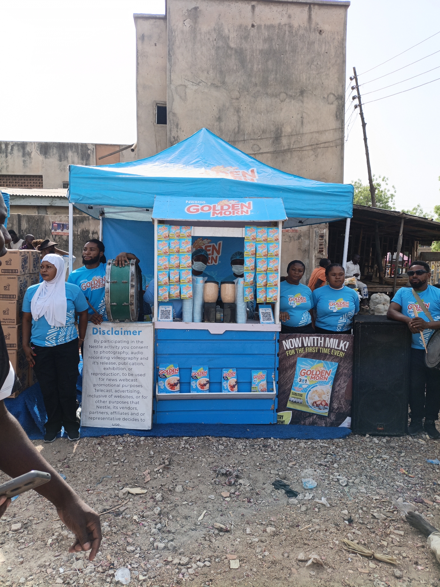 The image shows a promotional setup in the city of Kawaji, Nigeria. A blue-colored tent-like structure with "Nestle Golden Morn" branding is the centerpiece. Various Nestle Golden Morn products are displayed on the structure, including boxes of the cereal and a poster advertising "Golden Morn 3in1." Several individuals are present, some wearing blue t-shirts with the "Nestle Golden Morn" logo. One woman is wearing a white hijab. Some individuals are standing behind the promotional tent, while others are positioned on either side. A man on the right is holding drumsticks. A disclaimer regarding consent for photography and audio/video recording during the Nestle activity is displayed on a sign. The ground appears to be rocky and littered with debris. Building are behind the setup. A power pole is on the right.