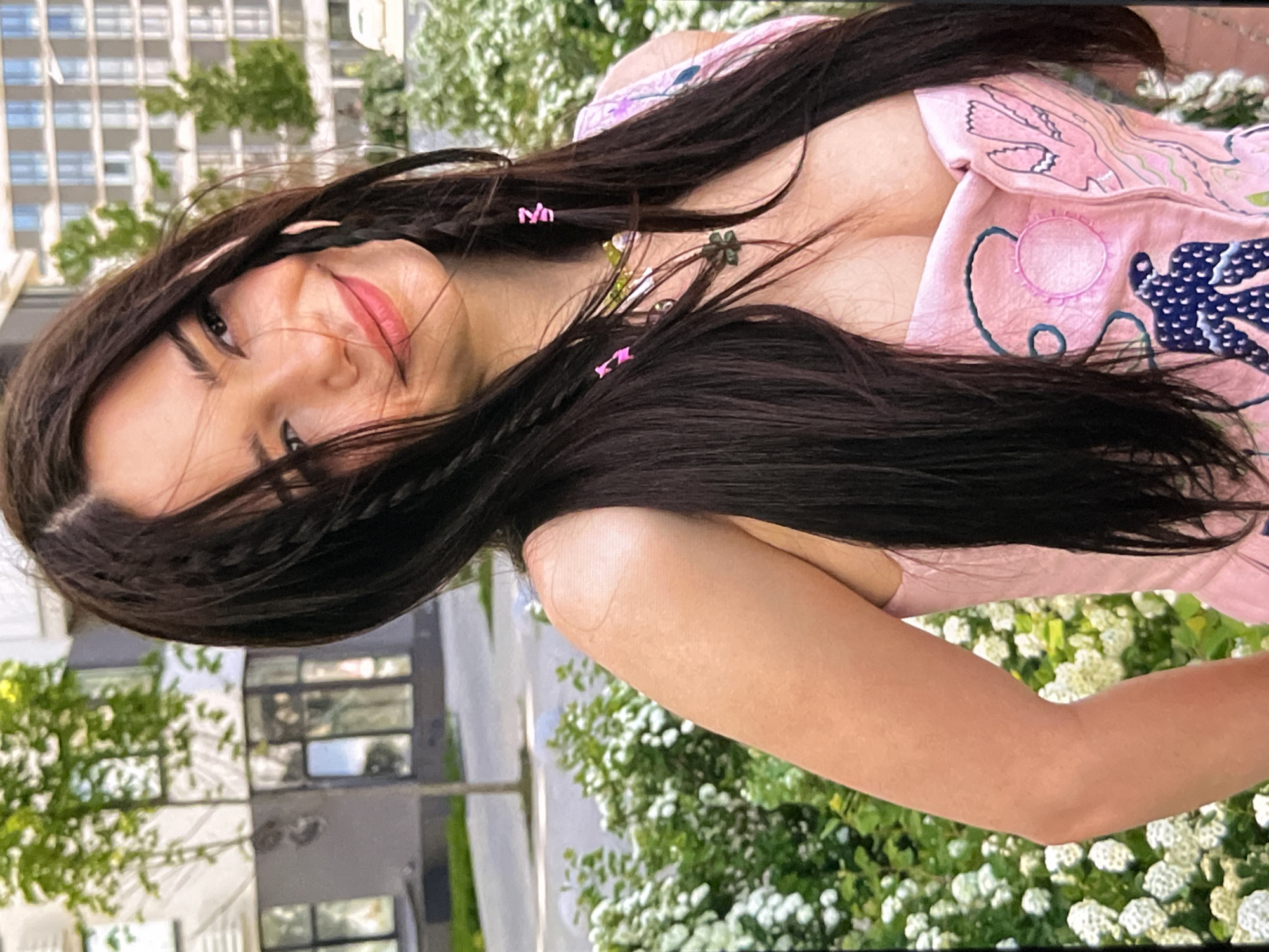 A close-up, eye-level, outdoor shot shows a young woman from the chest up. She has long, dark brown hair, styled with braids and small pink clips. Her head is tilted to the right, and she's looking to her right with a slight smile. She's wearing a pink bustier with intricate blue and white embroidered designs. In the background, there's a blurred green bush with white flowers, a sidewalk, and modern apartment buildings. The lighting suggests it's daytime.