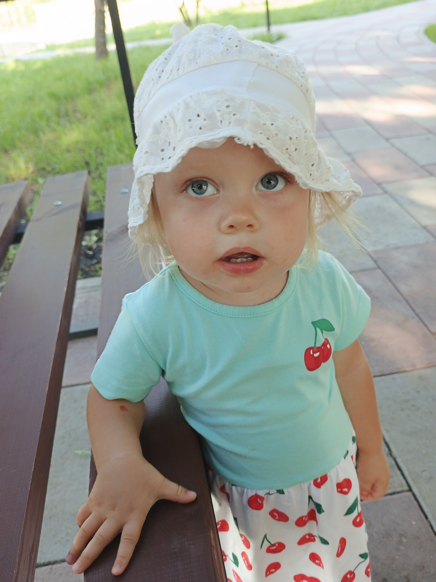 A blonde-haired toddler, wearing a white lace sun hat and a light blue t-shirt with a cherry print on the chest, stands in front of a brown wooden bench in an outdoor setting. The toddler has light skin and blue eyes. The t-shirt has a graphic of two red cherries with green stems. The toddler is also wearing a skirt or dress with a white background covered in a pattern of red cherries and green stems. The toddler's left hand is resting on the bench. Behind the bench, there is green grass and a paved walkway. The scene appears to be in a park or public outdoor space in Zlatoust, Russia.