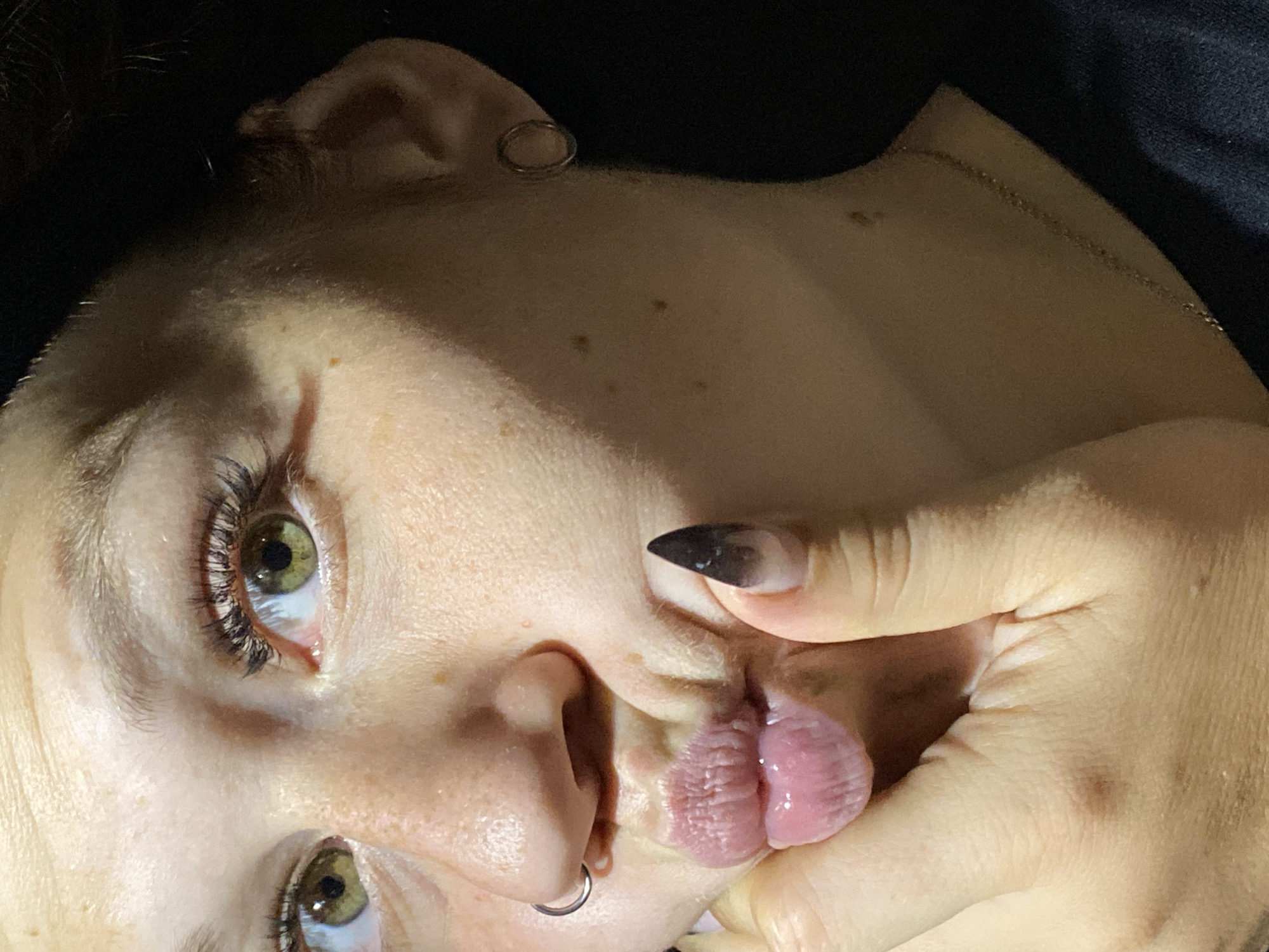 This is a close-up, chest-up, outdoor photograph of a young woman. She has fair skin, green eyes with long, dark eyelashes, and light brown eyebrows. Her lips are pink and slightly pursed, with her right hand holding her chin and lower lip. She is wearing a silver hoop earring in her left ear and a silver nose ring on the left side of her nose. Her fingernails are long and painted with a dark ombre effect. She is wearing a dark top and a thin gold chain necklace. The background is dark and out of focus, suggesting the photograph was taken at night or in low light conditions. There is no identifiable information about the location in Chaikovsky, Russia.