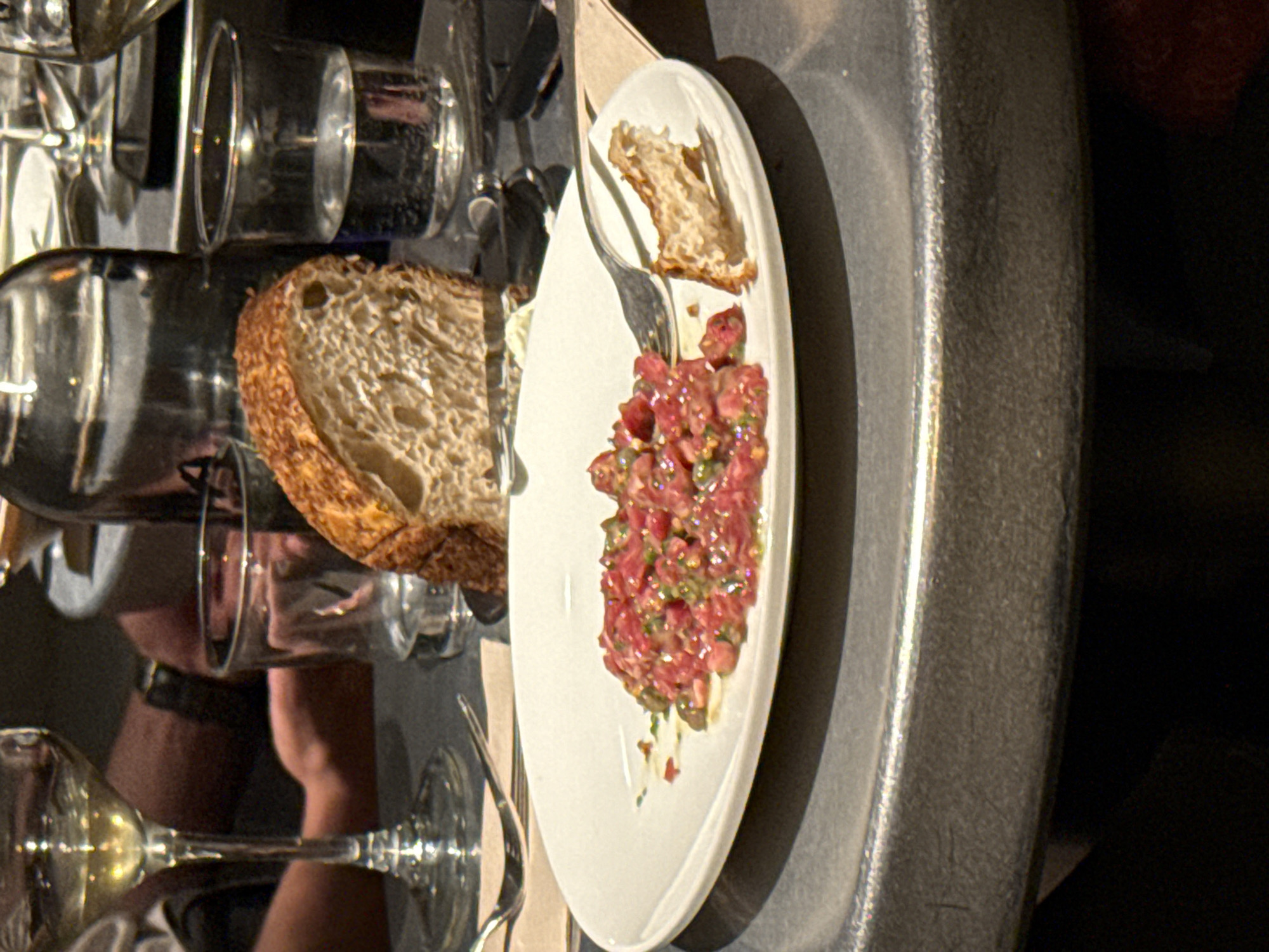 The image depicts a close-up of a table setting. A white plate with a serving of what appears to be steak tartare, accompanied by bread and a fork, occupies the center of the frame. In the background, there are multiple glasses, suggesting a dining or restaurant setting. An arm wearing a watch is also visible. The scene likely takes place at a restaurant or bar. It's impossible to definitively place this in the city of Madrid, Spain, with the information presented.