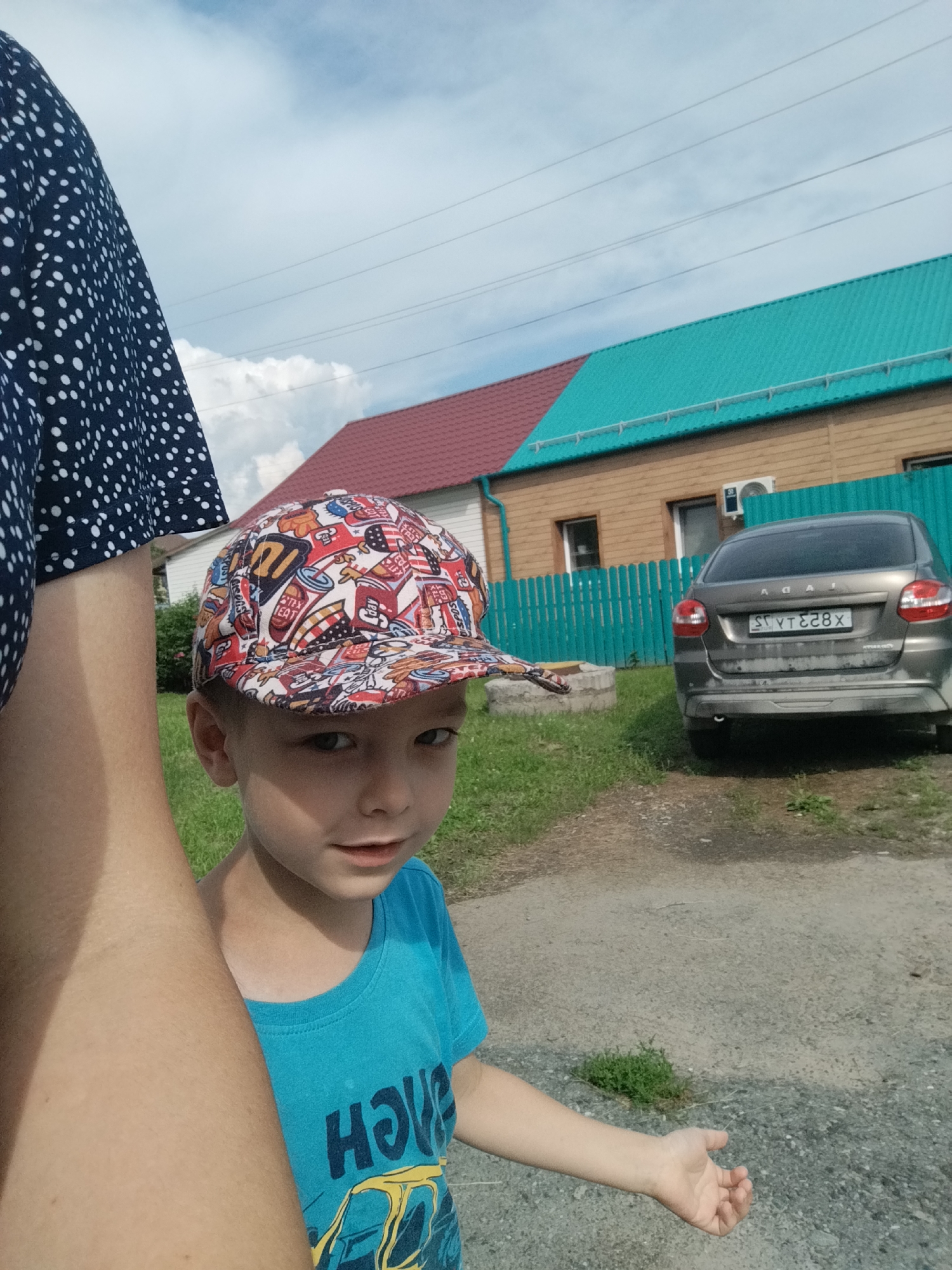 A medium shot captures a young boy in a blue t-shirt and a colorful, graphic-print baseball cap. He is looking towards the camera with a slight smile, his head tilted to the side. The boy's right arm is extended towards the viewer, his hand open and palm facing upwards, as if reaching for something. In the background, a grey sedan is parked in front of a wooden house with a teal-colored metal roof. A turquoise fence separates the yard from the house. The weather appears to be clear and sunny, suggesting a daytime setting. The scene is likely a residential area, possibly in Zavodoukovsk, Russia, given the context provided. The license plate of the car is partially visible, reading "ST YT828X". There are power lines running horizontally across the sky, and some green grass and a gravel path are visible in the foreground. The image has a casual, candid feel, as if it were a snapshot taken by a parent or guardian.