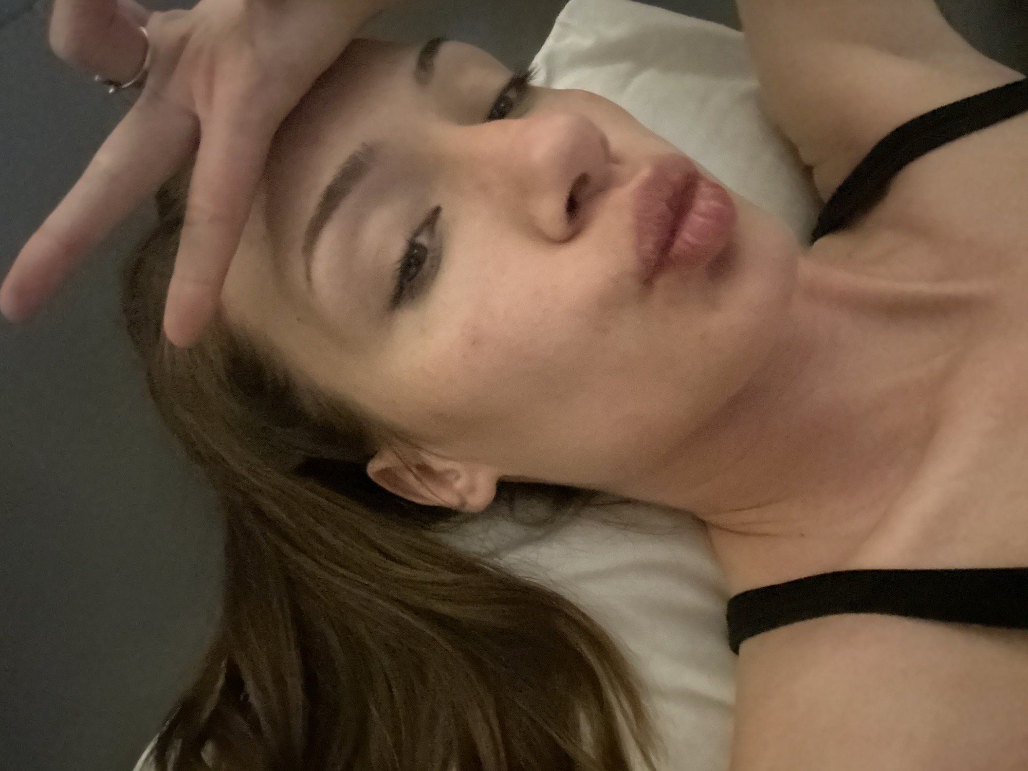 The image depicts a single female individual lying on a white pillow, presumably on a bed. The perspective is from above and slightly to the individual's right, suggesting a selfie. Her head is turned slightly to the left, and her gaze is directed towards the camera. Her lips are visibly puckered in a "kissing" gesture. Her right hand, wearing a silver-toned ring on the ring finger, is raised to the forehead area, forming a "peace sign" (V-sign) with the index and middle fingers extended. Long, dark brown hair is visible, flowing across the pillow and a grey surface. She is wearing a black strap on her left shoulder, consistent with a bra or tank top. The skin tone is fair, with minor facial blemishes visible. The environment appears to be an indoor residential space. Visual details within the image do not provide specific identifiers for a location in Varna, Bulgaria.