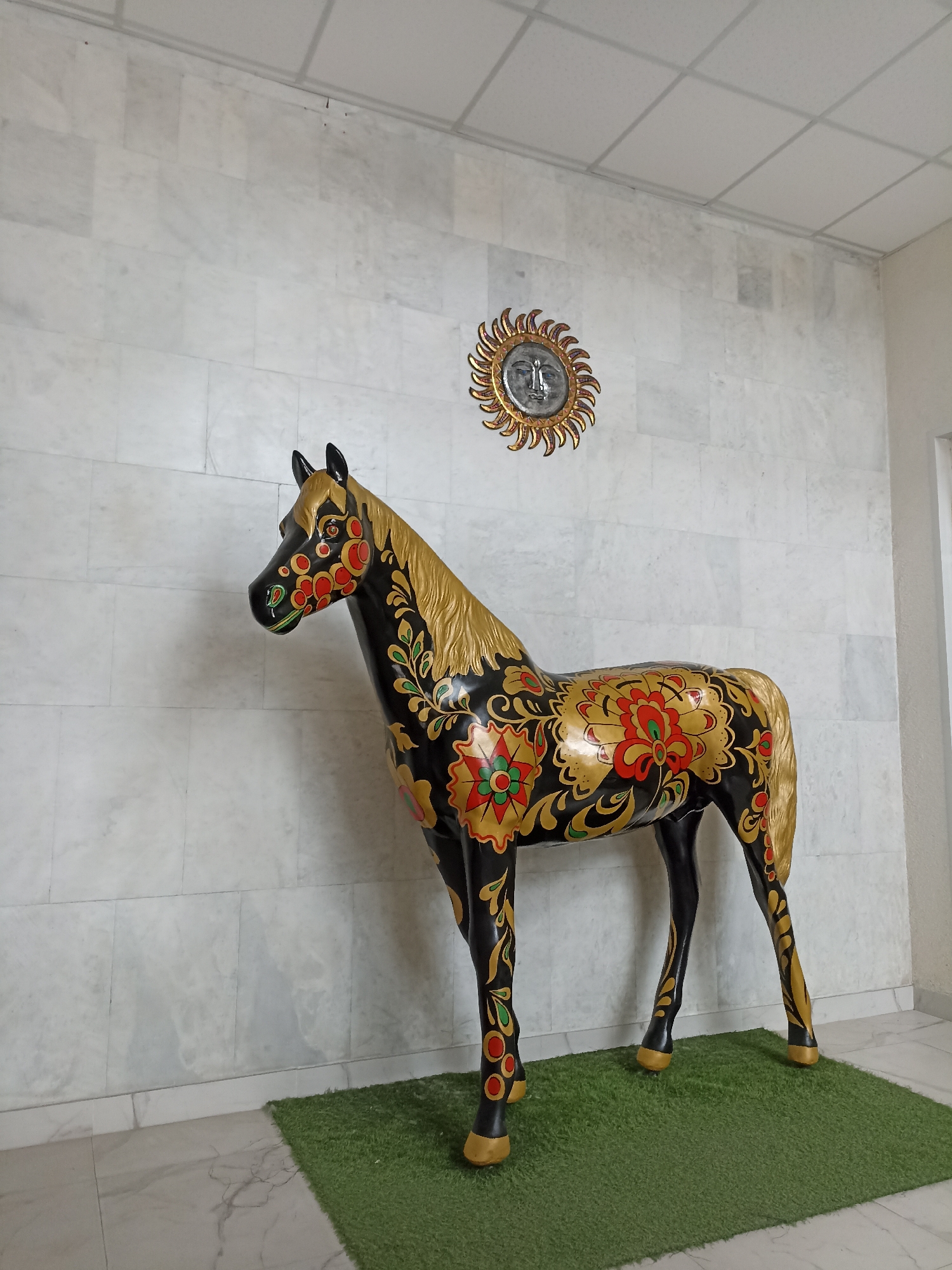The image shows a life-sized statue of a black horse with ornate gold, red, and green floral patterns. The horse is standing on a small patch of green artificial turf against a white marble tiled wall. Above and to the right of the horse, a decorative sun clock with a human face and radiating rays is mounted on the wall. The floor is tiled with a marble pattern. The overall scene appears to be an indoor public space, possibly a lobby or a museum exhibit. There is no indication of the specific location being Novosibirsk, Russia.