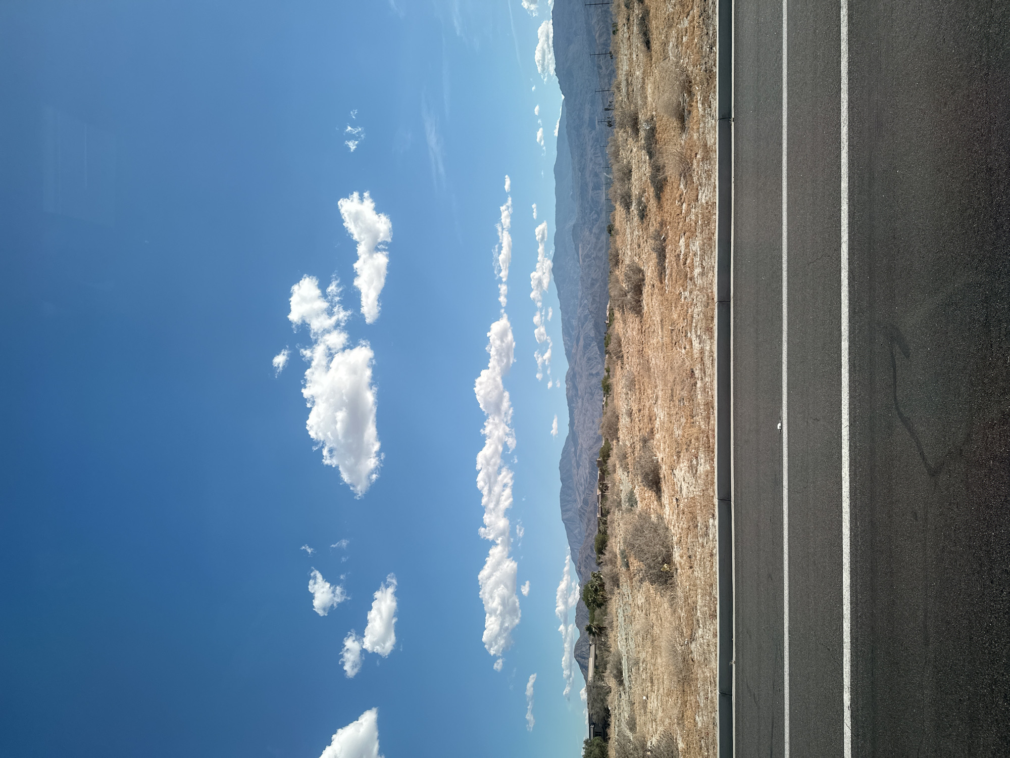 The image depicts a landscape in an arid environment. A clear blue sky with scattered cumulus clouds is visible above a mountain range in the distance. Adjacent to the dry, scrubby terrain is a paved road with multiple lanes separated by white lines. The perspective suggests a viewpoint from an elevated position, likely a moving vehicle. No people are visible in the frame. The location is unknown, but the landscape indicates an arid or desert region, possibly in a mountain area, given the vegetation and mountain range.