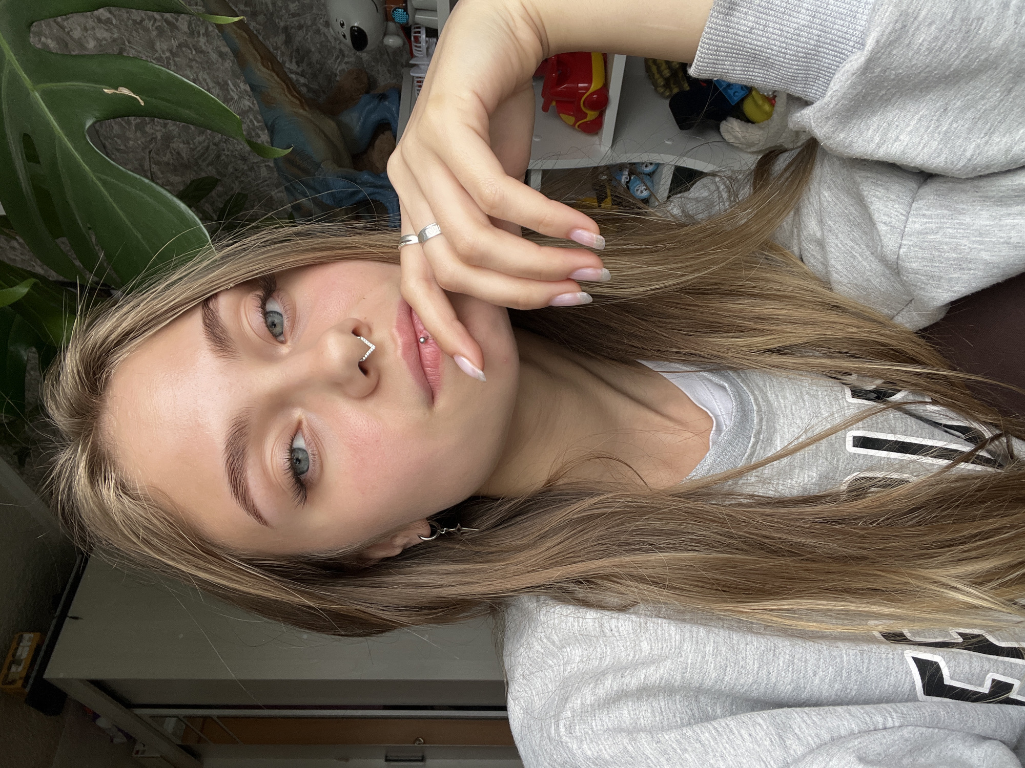A young woman with long blonde hair is facing the camera. She has light skin, blue eyes, and pierced nose and lips. She is wearing a gray sweatshirt. Her left hand is raised to her face, adorned with a silver ring. A large green plant is visible on the left side of the frame. In the background, there's a shelf cluttered with miscellaneous items, including toys and clothing. The location is unspecified and there is no evidence to suggest the location is Фоки, Russia.