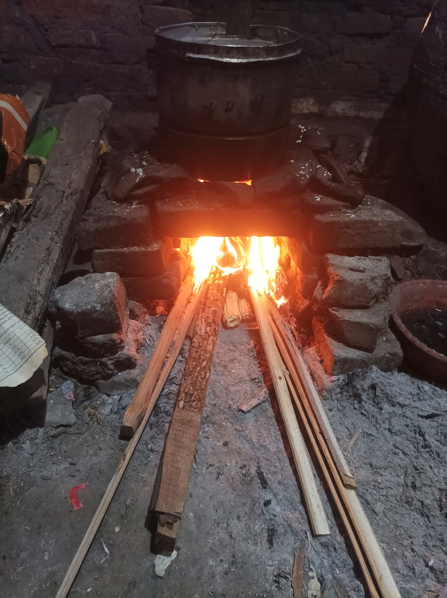 A rustic outdoor cooking scene is depicted in Canga'an, Indonesia. A fire burns intensely within a stone structure, fueling a large metal pot positioned above it. The area is littered with ash, firewood, and miscellaneous debris, suggesting a practical and functional setup for preparing food. The lighting is dim, with the flames providing the primary illumination, casting an orange glow on the surrounding elements.