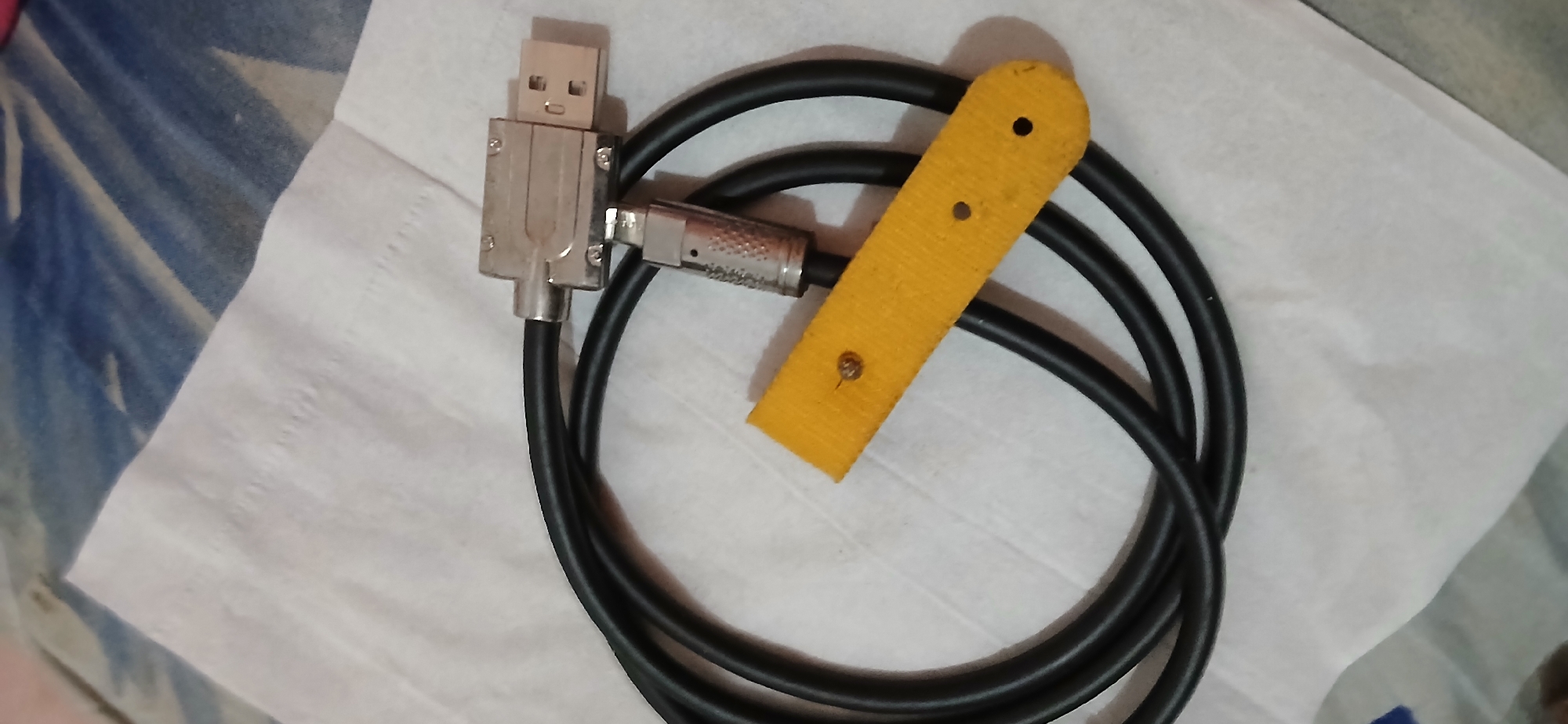 The image shows a close-up, overhead view of a black cable coiled on a white textured surface, likely a paper tissue. The cable has two connectors. One is a silver-toned USB-A male connector, angled at 90 degrees from the cable. Attached to this connector is a smaller, cylindrical silver connector. A yellow fabric strip, possibly a cable tie or strap, with two small holes and a tiny screw or rivet is positioned under the coiled cable. The background is blurred, showing a patterned surface with blue and white tones, suggestive of fabric. The image was taken in Samarinda, Indonesia.