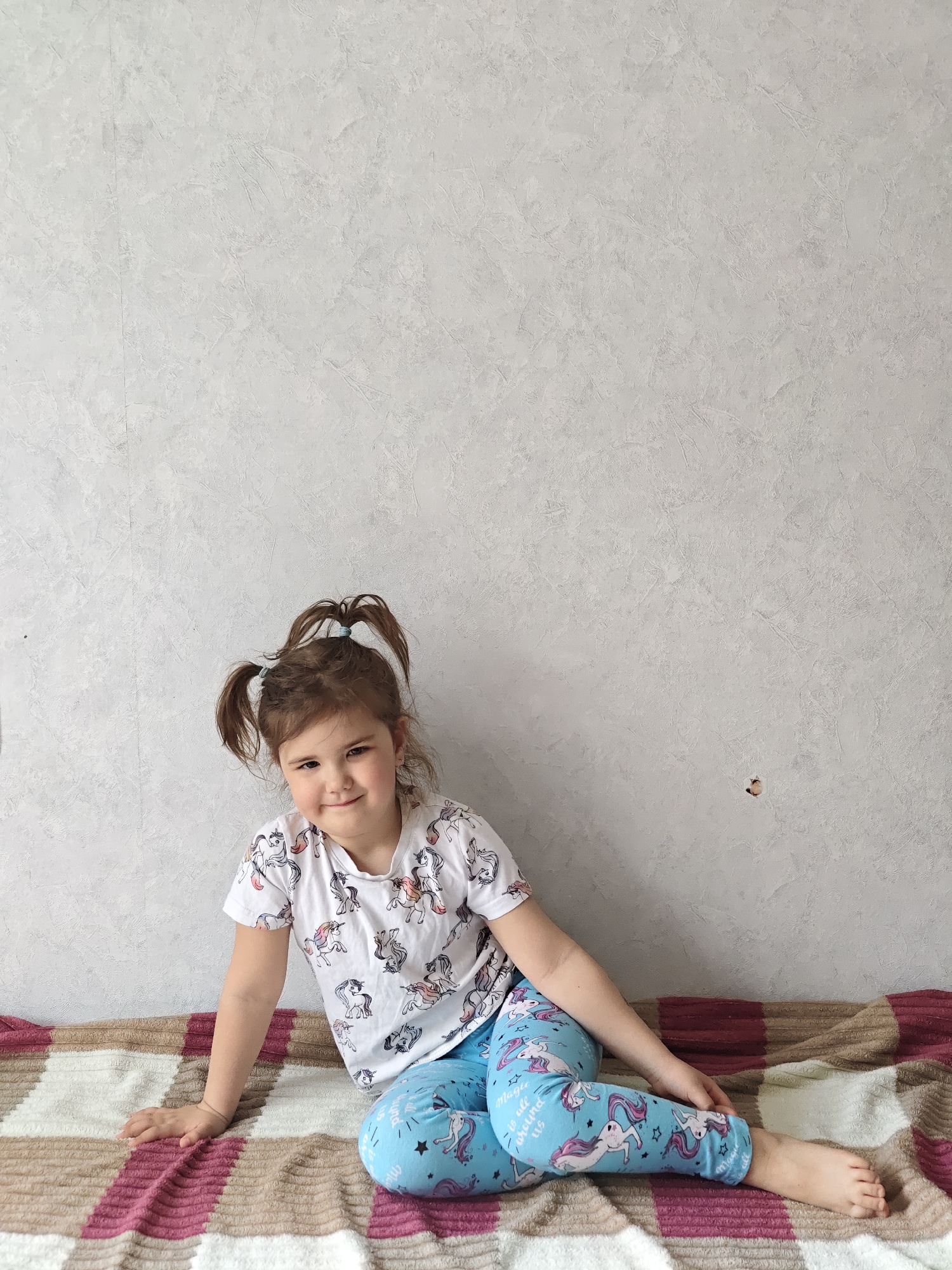 A young girl with brown hair in pigtails sits on a patterned blanket in front of a textured gray wall. She is wearing a white t-shirt with a unicorn print and light blue unicorn leggings. Her left hand is resting on the blanket and her right hand is near her right leg. The location is in the city of Chaikovsky, Russia.