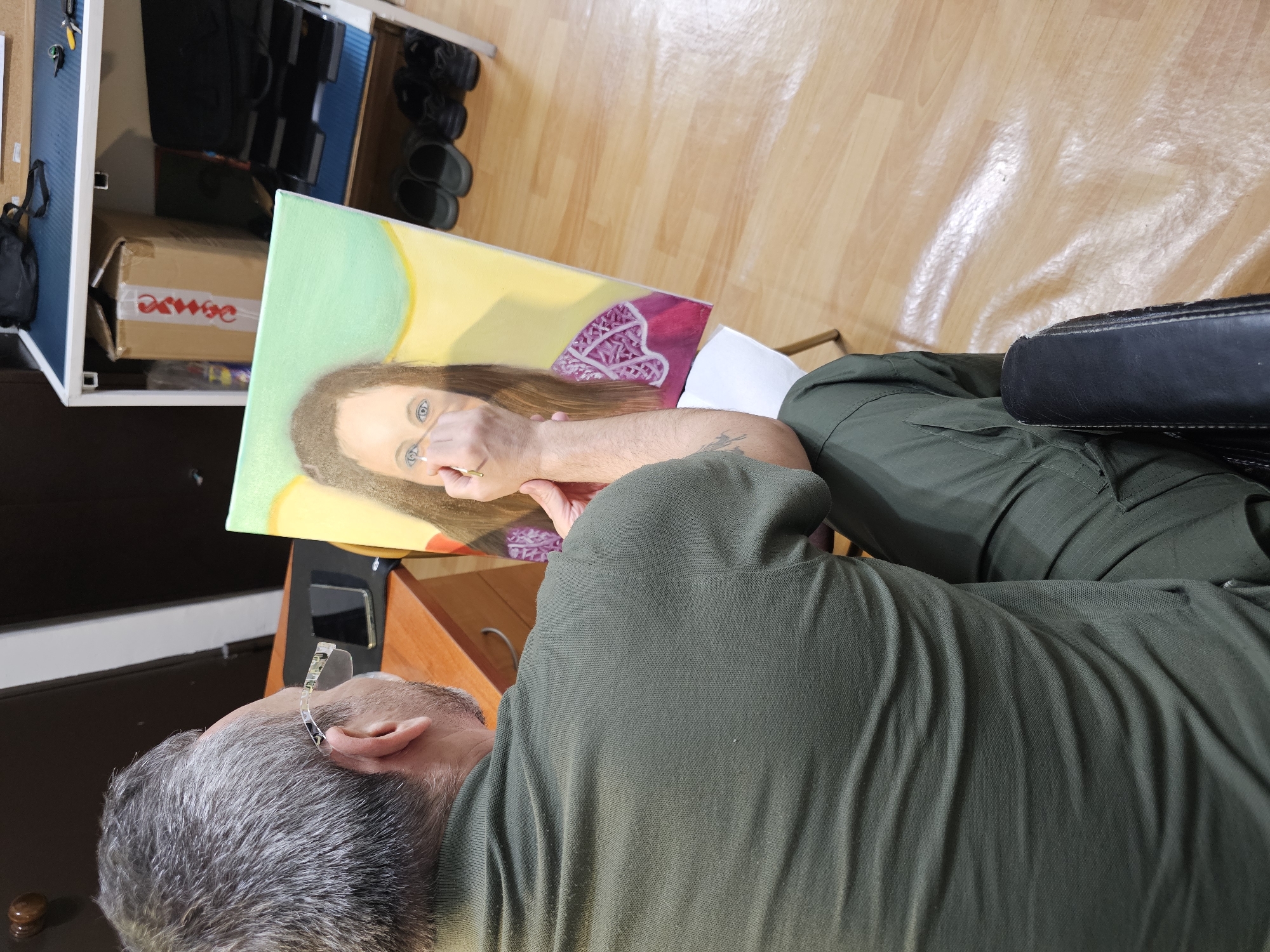 An overhead shot captures a man engaged in painting a portrait indoors, likely in an art studio or home in Moscow, Russia. The man, with graying hair and a beard, is seen from behind and above, wearing a dark green polo shirt and matching pants. Glasses are perched on his head.
He is seated in a dark chair, with only an armrest visible, and is intently focused on a canvas positioned on an easel in front of him. His right arm is extended, holding a thin paintbrush, which he uses to add fine details to the subject's right eye. The canvas displays a vibrant portrait of a woman with long, flowing brown hair and striking blue eyes. Her right hand is painted in a distinctive pose, held up and partially obscuring her nose and mouth. The background of the portrait is abstract, featuring sections of light green, yellow, and a magenta area with a complex geometric pattern.
The setting includes a light wooden floor that reflects light, suggesting daytime. In the background, a white shelving unit is visible, holding a black bag, black shoes or slippers, and a brown cardboard box. The text "ДЕЛО" (reversed) is visible on the side of the box.