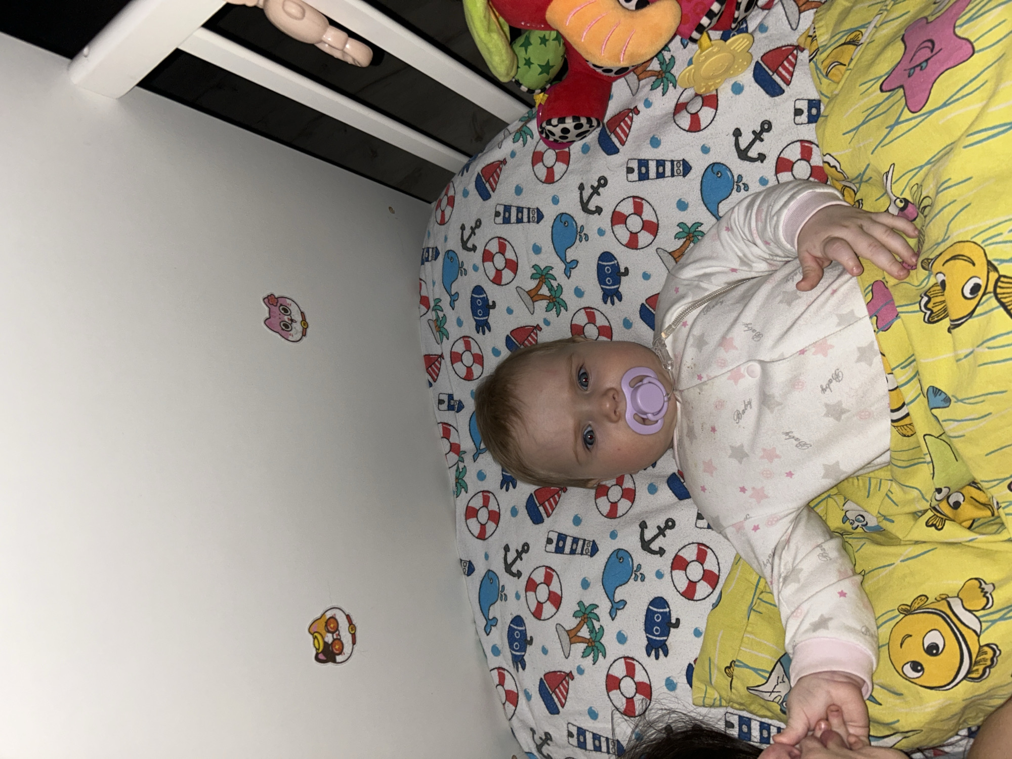 An overhead indoor shot captures a baby lying in a crib in Rusalevka, Russia, with an adult's hand gently holding theirs. The baby has light hair and blue eyes, is wearing a white sleeper adorned with small pink stars and the repeated word "Baby," and has a purple pacifier in their mouth. They look directly towards the viewer.
The crib features white bars, and above the baby, a colorful mobile with plush toys is partially visible. The baby is resting on a fitted sheet with a playful nautical pattern, including blue whales, red and white lifebuoys, anchors, palm trees, boats, and submarine-like figures. A bright yellow blanket, decorated with cartoon fish resembling clownfish and pink starfish, covers a portion of the baby.
The white wall beside the crib is decorated with two small, colorful stickers: one depicting a pink owl-like character and another featuring a brown and orange owl-like character. The lighting suggests an indoor environment, possibly a nursery or bedroom, under soft or ambient light conditions.