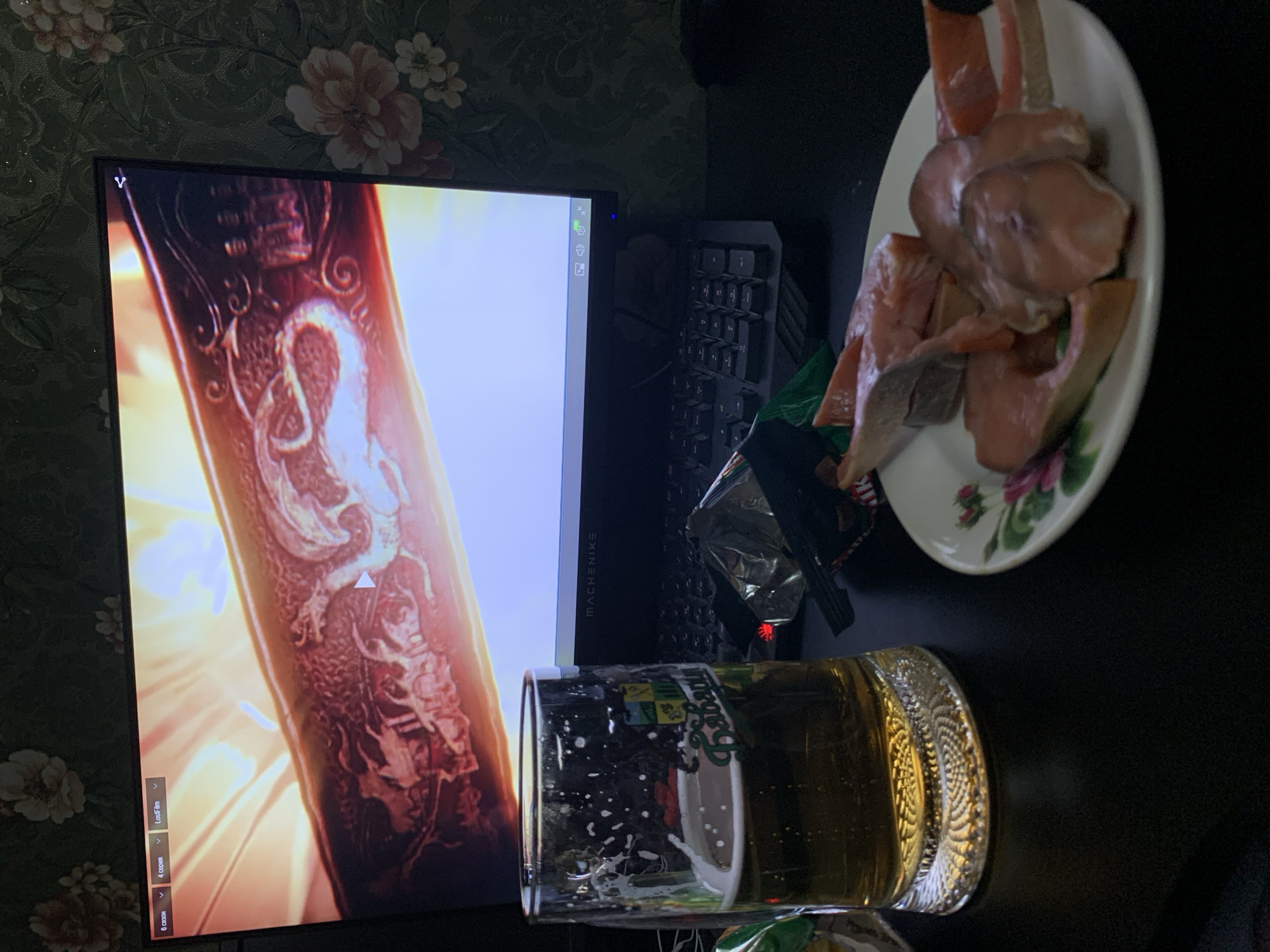 The image depicts a dark tabletop setup featuring a beer mug, a plate of raw fish slices, and a computer monitor displaying what appears to be a scene with dragons. The monitor is a MACHENIKE brand, and the screen displays a menu with options for "6 сезон" (Season 6), "4 серия" (Episode 4), and "LostFilm". The background of the monitor shows a close-up of a metallic object with intricate dragon engravings, illuminated by bright light. To the left of the monitor is a beer mug filled with a golden beverage and foam, with a label on the glass reading "Балтика" (Baltika). To the right of the beer mug and in front of the monitor is a crumpled silver foil packet, likely packaging for snacks. The plate in the foreground contains several raw slices of pinkish-orange fish, possibly salmon. The tabletop is black and reflective. The background of the image features a wall with a floral wallpaper pattern in shades of green and peach. The scene is lit from the monitor, creating reflections on the tabletop and the beer mug. The overall impression is a casual, indoor setting, potentially a person's workspace or entertainment area. There are no people visible in the image. The location is stated as Desyatikhatka, Russia.