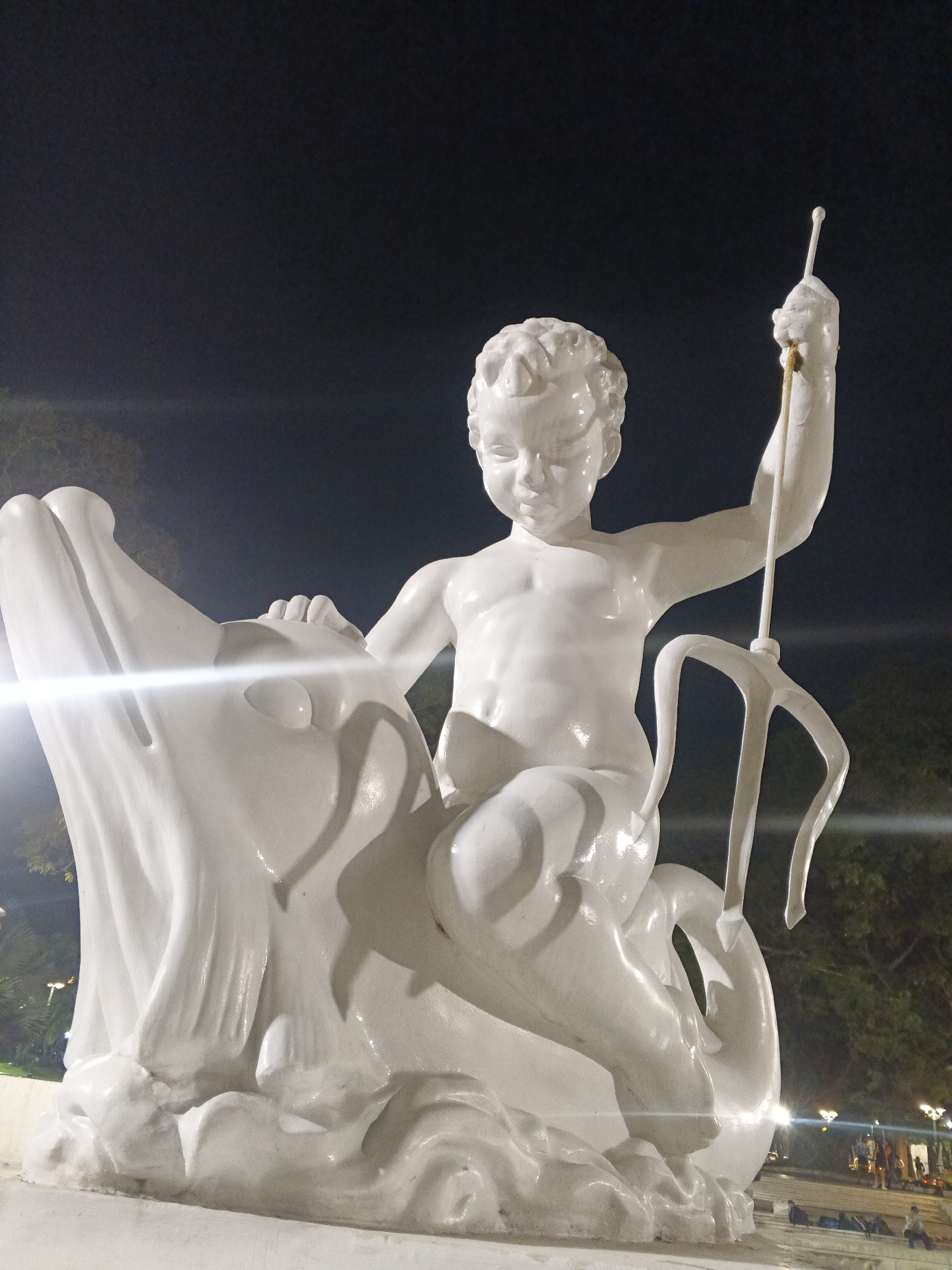 The image shows a white sculpture of a child riding a dolphin, holding a trident. The sculpture is illuminated at night, with a dark sky in the background and some trees and lights visible in the distance. The sculpture is located in the Plaza del Chupones in Caracas, Venezuela. There are some people sitting on the ground in the background, and bags are placed near them. The lighting from street lamps and possibly spotlights creates strong highlights and shadows on the sculpture, emphasizing its form and texture.