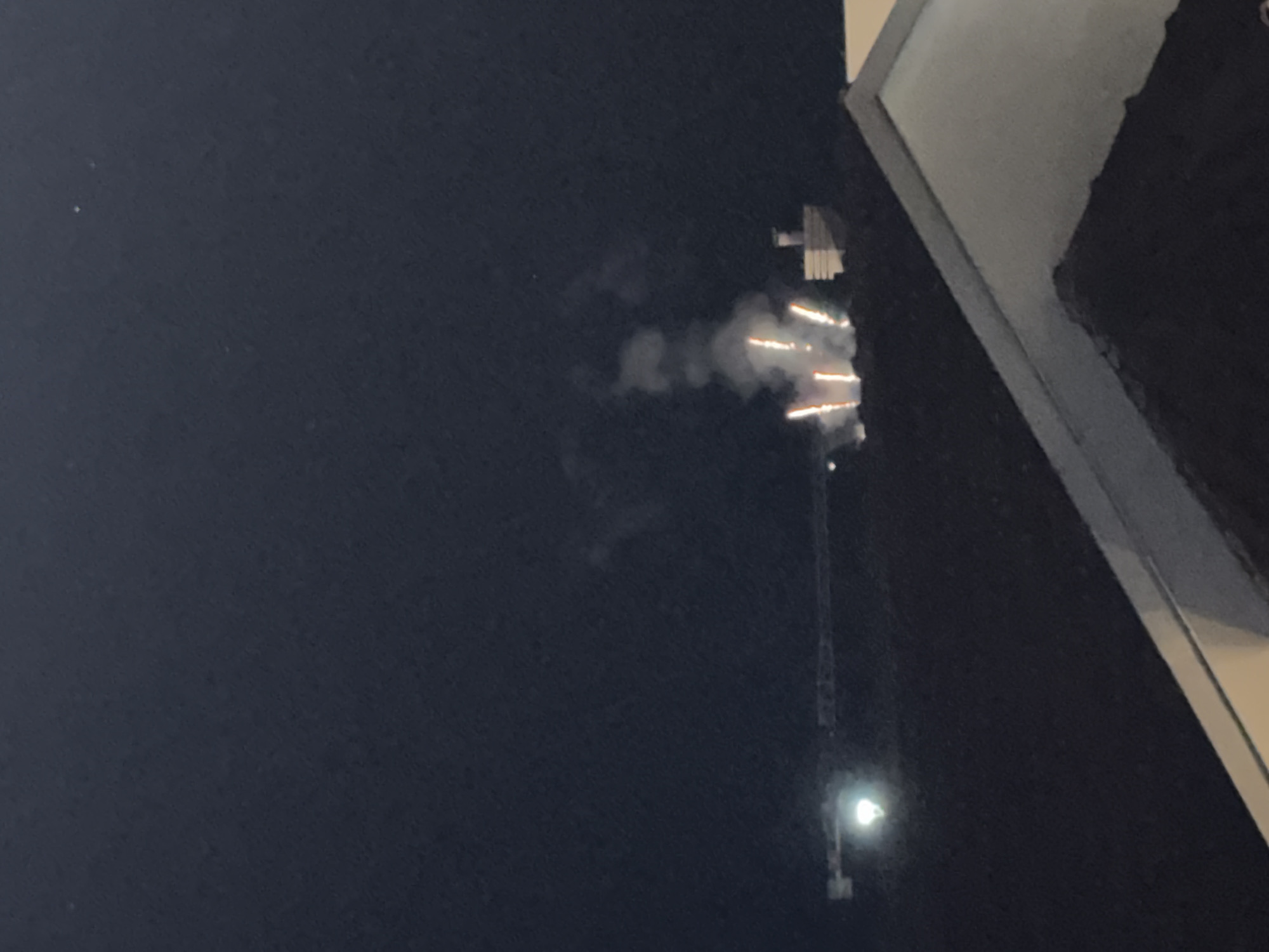 The image depicts a nighttime scene with a dark sky. In the mid-ground, there are several bright, vertical streaks of light that appear to be fireworks. These are accompanied by plumes of smoke. To the right of the fireworks, there is a distinct building with a layered, angular roof structure. Further to the left, a long, horizontal structure, possibly a crane, is visible with a bright light at its left end. In the foreground, the roofline of a building is visible, obscuring the lower portion of the scene. The image quality is somewhat grainy due to low light conditions. The context provided suggests this scene is in the city of Trap, Montenegro, though the visual information in the image itself does not directly confirm this specific location.