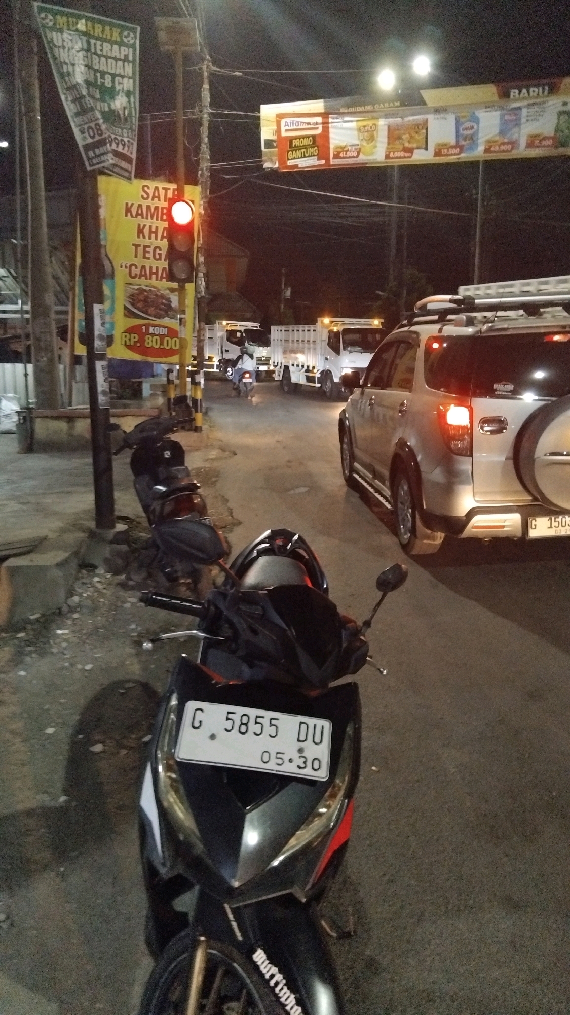 The image captures a street scene at night in Tegal, Indonesia, as evidenced by the Indonesian language on the advertisements and license plates. In the foreground, a black motorcycle is parked, with its license plate clearly visible as "G 5855 DU 05.30". To the left of the motorcycle, a traffic light is illuminated red. Behind the motorcycle, several other parked motorcycles are visible. The street is lined with poles holding various advertisements, including one for "Alfainur" and another promoting "Sate Kambing". Several white delivery trucks are driving on the road, indicating a commercial or industrial area. A silver SUV is also present on the road, with its brake lights illuminated. The overall environment appears to be an urban street with moderate traffic activity.