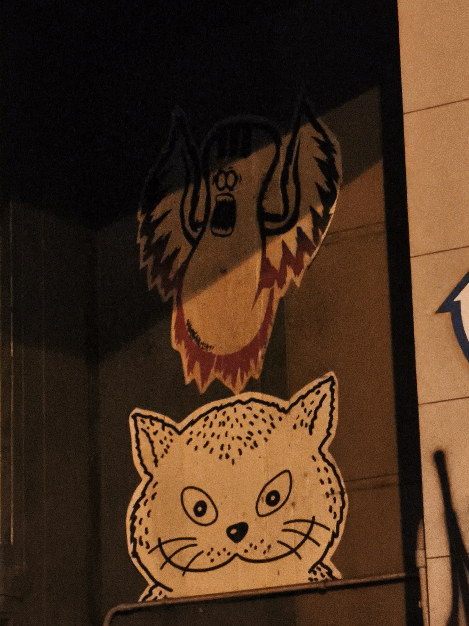A night shot shows two cartoonish paper decorations on a storefront window in Barcelona, Spain. The top image is a yellow and red figure with spiked edges around the face, wide eyes and an open mouth, possibly suggesting fear or surprise. The bottom image is a cat's face, also yellow, with black outlines, dots, and whiskers. The background is dark, with a light wall to the right that includes a blue and white arrow. The overall impression is whimsical and slightly eerie, especially in the dark setting.