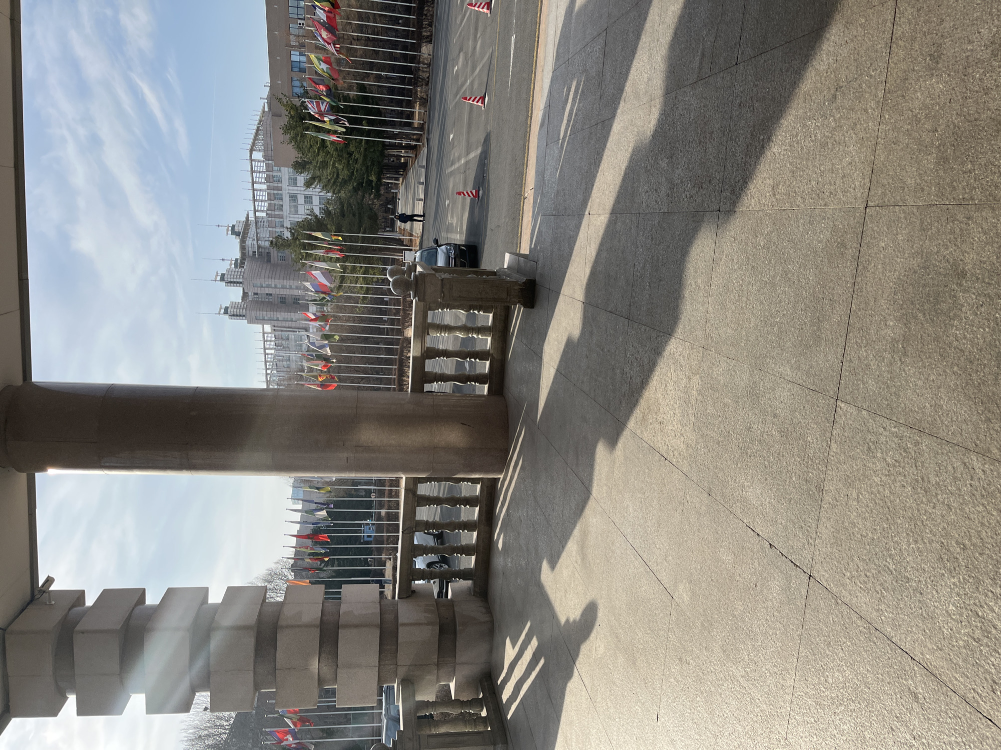 The image shows a partial view of a stone structure, likely a building or monument, in Changchun City, China. The view is from an elevated position, looking out onto a plaza or street. There are several flagpoles with international flags visible, suggesting the location might be related to a government building or international organization. A black car is parked in the street. The ground is paved with large stone tiles, and shadows of the building structure cast over it. The sky is blue with some clouds. There is a single person in the background and orange safety cones on the road.