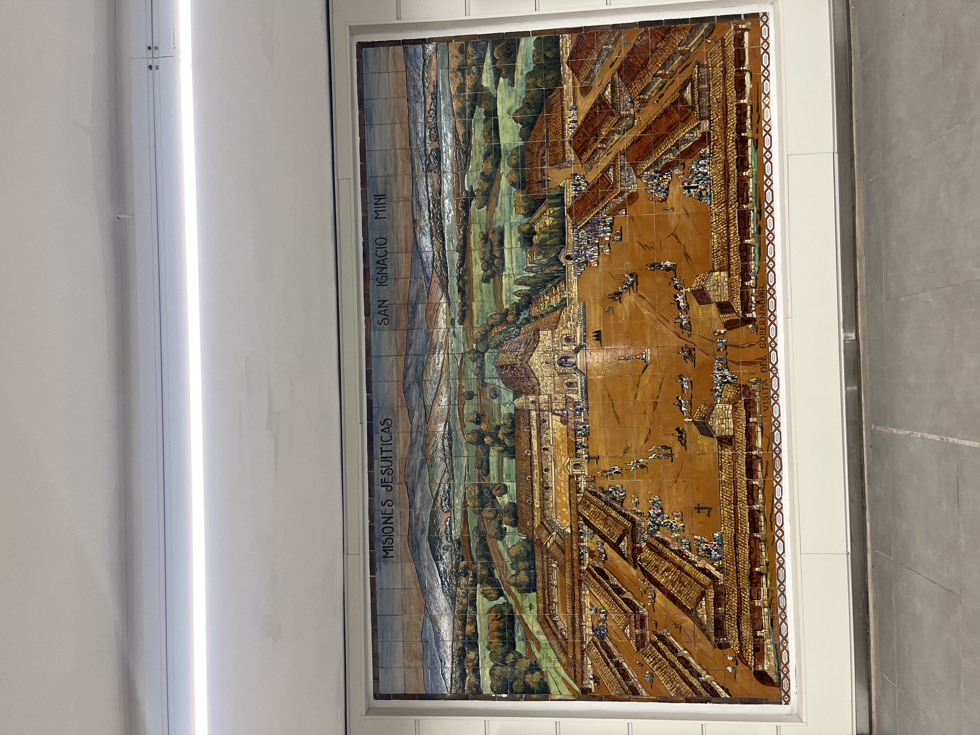 A colorfully tiled mural adorns a wall in what appears to be a building interior. The mural depicts a landscape scene with what appears to be buildings and figures, possibly a historical site. Text on the mural reads "MISIONES JESUITICAS SAN IGNACIO MINI." The image is likely taken in Buenos Aires, Argentina, as that city contains a metro station named "San Ignacio," which features tiled murals depicting Jesuit Missions. The area around the mural is minimally decorated, with a fluorescent light visible on the adjacent wall.