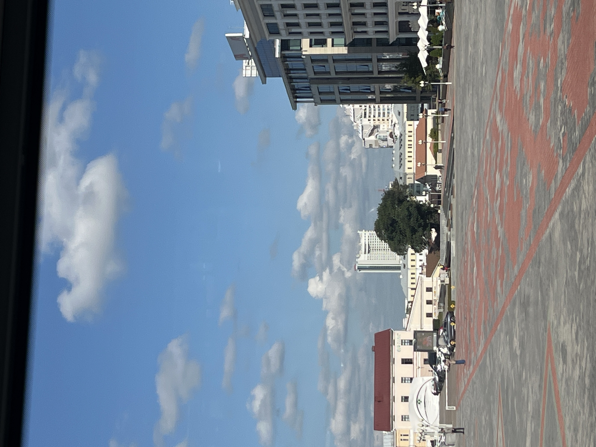 The image depicts a bright, sunny day in a public square in Minsk, Belarus. The square is paved with grey and red interlocking stones, forming a patterned surface. In the background, several buildings of varying architectural styles are visible, including a modern glass-fronted office building on the right, a red-roofed building with multiple windows in the center-left, and a tall, white skyscraper in the distance. The sky is a clear blue with scattered white clouds. Several people are present in the square, walking or standing. Cars are parked in the background near some of the buildings. The scene suggests a bustling urban environment under a pleasant sky.