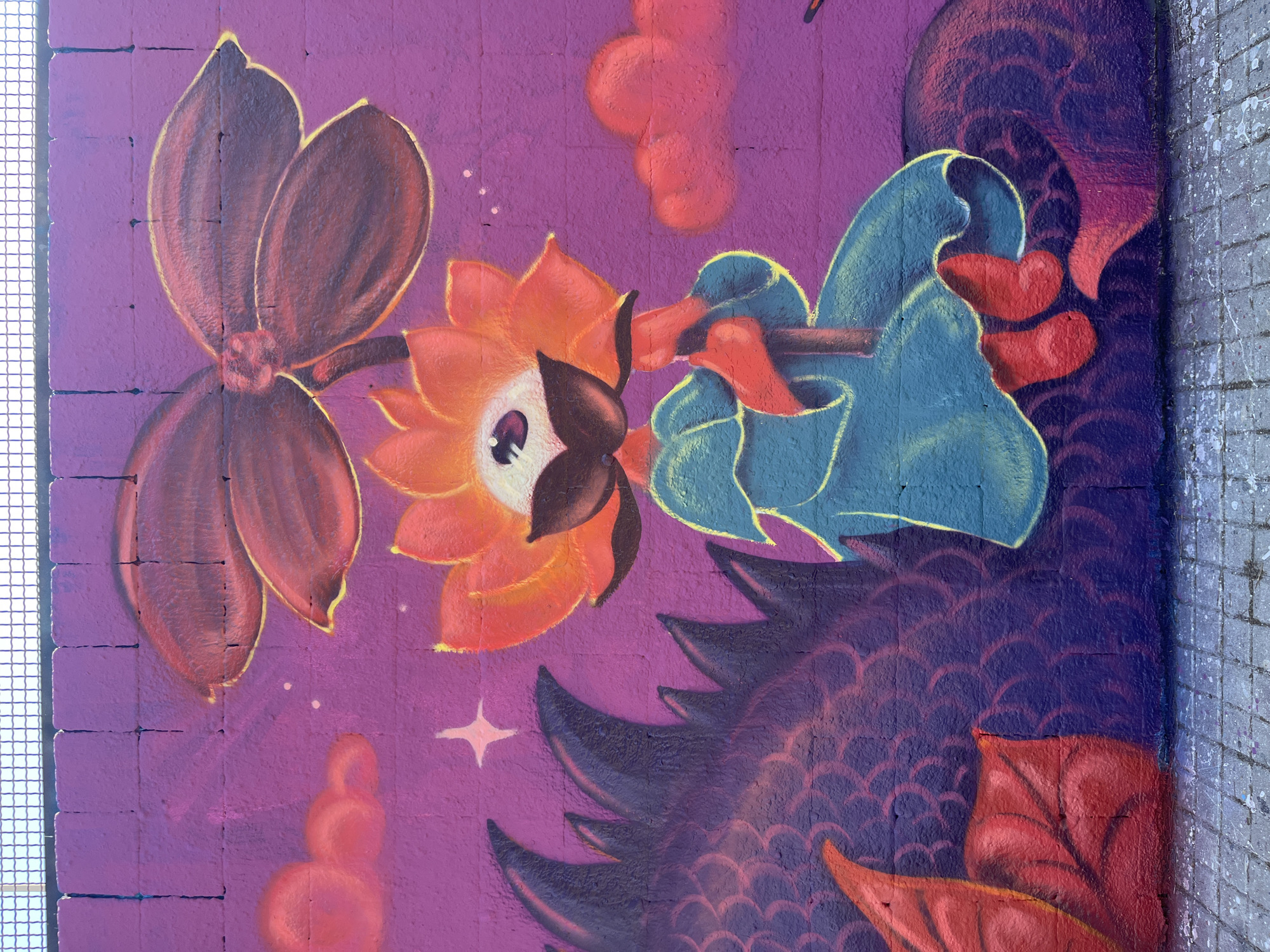 This media file captures a vibrant and detailed section of a mural, likely painted on a concrete block wall in Barcelona, Spain. The artwork features a whimsical, anthropomorphic flower character at its center, rendered primarily in shades of orange and red with subtle yellow highlights. This character has a large, stylized eye with a white iris and a black pupil, and dark, petal-like structures framing its "face."
The flower character is depicted holding a dark, cylindrical pipe or flute-like object. On the other end, a blue, irregularly shaped element, resembling either a stylized hand, wing, or fabric, also grasps the pipe. Below this blue element, two distinct red heart shapes are visible, adding to the playful and endearing quality of the artwork.
The background is a rich, mottled purple, against which smaller, puffy red cloud-like shapes and delicate white starbursts are scattered, suggesting a fantastical sky or dreamscape. Towards the bottom and right of the image, a large, dark purple form with intricate, lighter purple and red scale-like patterns is visible, suggesting part of a mythical creature such as a dragon or fish. Below this, prominent red leaf-like or wing-like structures emerge.
The wall itself shows texture, with visible mortar lines of concrete blocks, and a grid-like white fence or grate runs along the left edge of the mural. The right side shows a rougher, grey wall with white paint splatters. The overall style is cartoonish and illustrative, utilizing bold outlines and a vivid color palette. No text is visible in the image.