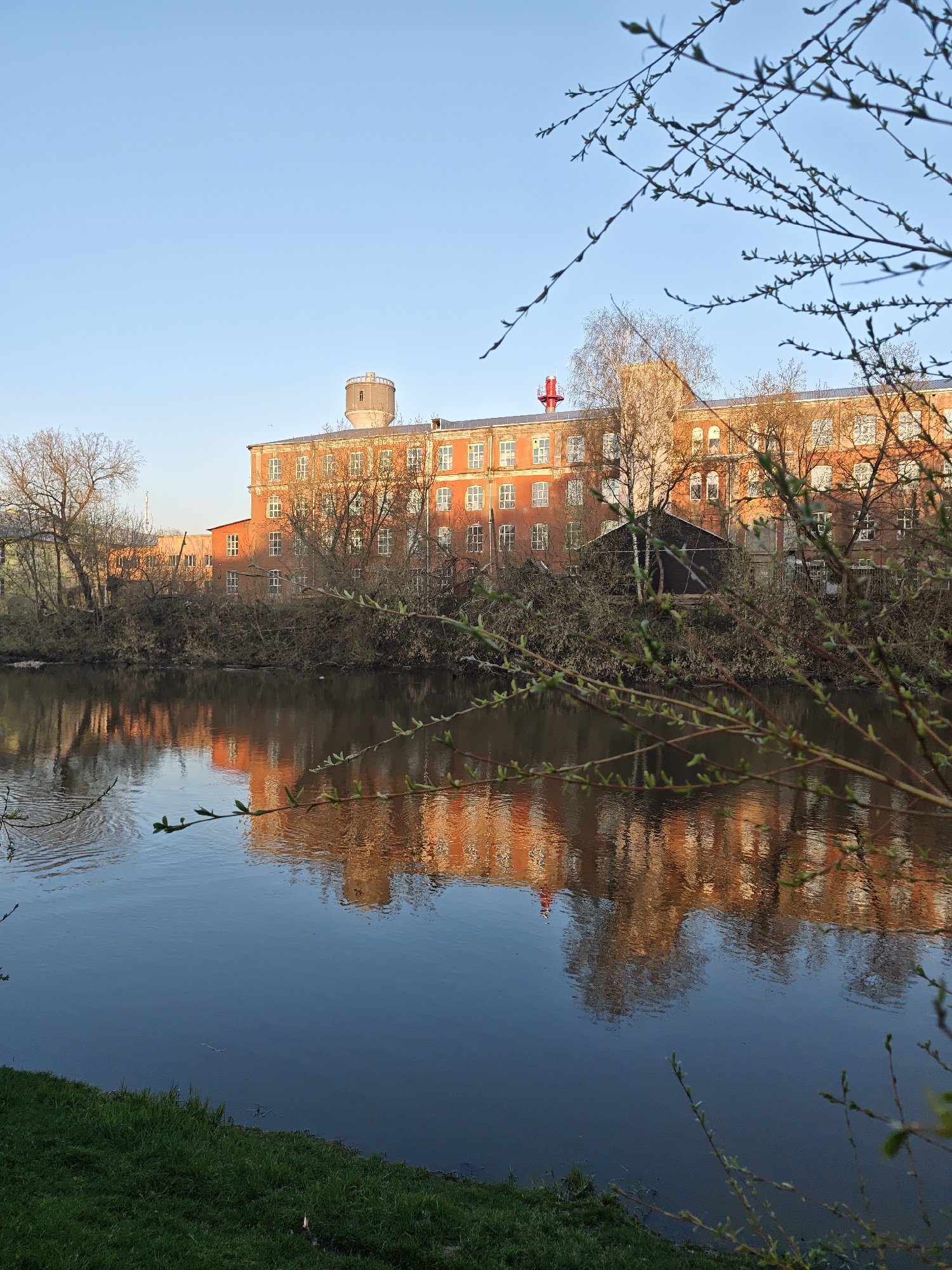 The image depicts a waterfront scene in Noginsk, Russia. In the background is a long, multi-story red brick building with numerous windows, likely a factory or industrial building. A cylindrical tower sits atop one section of the building, and a red structure resembling a cross or weather vane is visible on another section. Bare trees and shrubbery line the waterfront. The building and trees are reflected in the calm water of the foreground, which appears to be a river or pond. A small patch of green grass is seen along the bottom edge of the frame. In the foreground, branches with emerging leaves frame the scene, adding depth and indicating the early spring season. The sky is clear and blue, suggesting a sunny day.