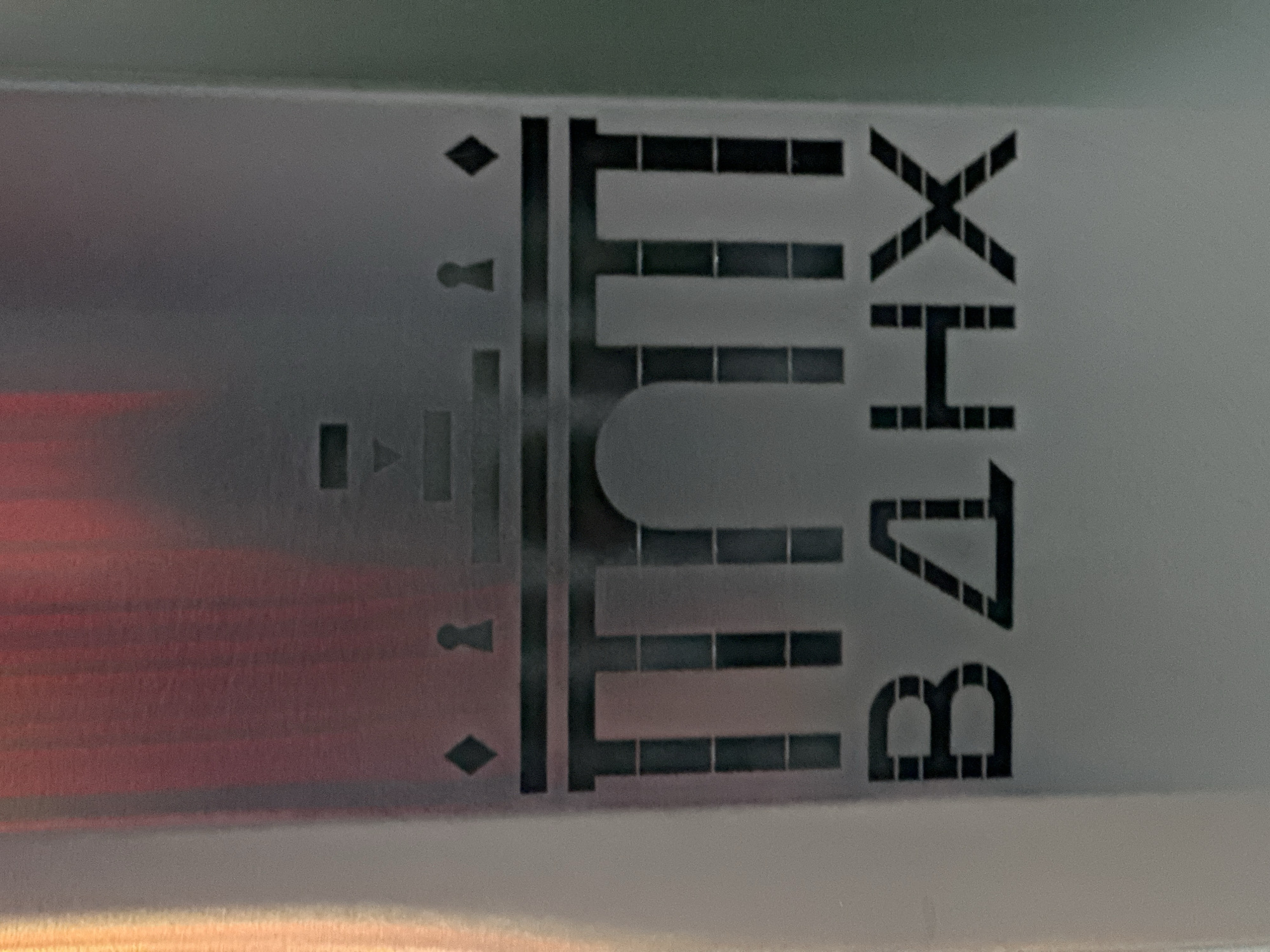 The image displays a close-up view of stenciled alphanumeric characters and symbols on a light-colored, smooth surface, possibly metallic or plastic. The image appears to be rotated 90 degrees counter-clockwise.
When viewed upright, the visible text and symbols extend horizontally across the frame. To the left of the main text, a sequence of black, solid shapes is arranged vertically: a diamond, followed by a keyhole-like icon, a small rectangle, another keyhole-like icon, and another diamond.
The main inscription, constructed from individual rectangular black blocks, reads "TUNTI B4HX". The "T" is a solid vertical block with a top bar. The "U" is characterized by a rounded bottom formed by blocks. The "N" and "I" are formed by standard block arrangements. The "B" is composed of two distinct rounded arch shapes made from blocks. The "4" is rendered as a triangular shape, the "H" is a standard block-letter H, and the "X" is formed by diagonal blocks.
The surface itself is reflective, showing a reddish-orange glow in the bottom-left portion, which suggests an external light source or colored reflection. The upper background shows a subtle greenish tint. The precision of the stenciling and the alphanumeric code indicate that this is likely a marking on an industrial product, an electronic device, or a piece of equipment. There are no human subjects or any activity taking place in the image. The setting appears to be an indoor environment due to the controlled lighting and clean surface.