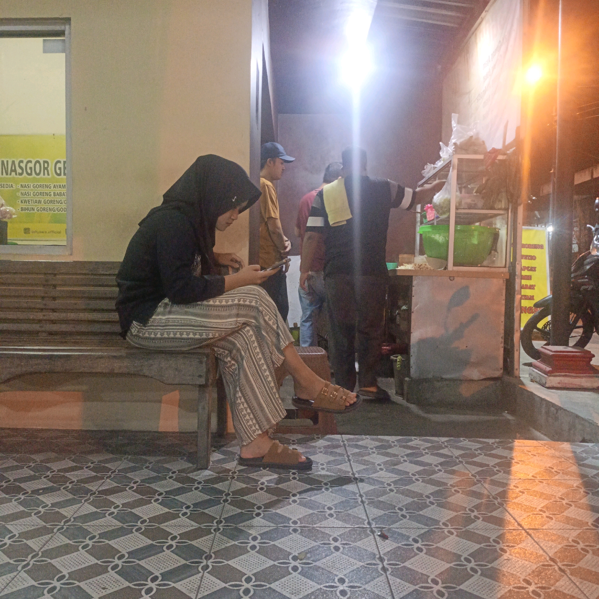 The image depicts an outdoor scene at night, possibly a street food stall or eatery, located in Salatiga, Indonesia. In the foreground, a woman wearing a black hijab and patterned pants is seated on a wooden bench, engrossed in her mobile phone. She is wearing brown sandals. The floor is tiled with a geometric pattern.
In the background, a food stall is visible, with shelves displaying ingredients and cooking utensils. A bright light illuminates the area. Three men are present near the food stall. One man, wearing a yellow t-shirt and a blue baseball cap, stands facing away from the camera. Another man, wearing a red shirt, is partially visible. A third man, in a black shirt with white stripes on the sleeves, is attending to the food stall, with a yellow towel draped over his shoulder. A motorcycle is parked to the right of the food stall. A sign with text in Indonesian is visible on the left side of the image, indicating food items like "NASGOR GE," "NASI GORENG AYAM," and "NASI GORENG BABAT." The overall ambiance suggests a casual dining or takeaway spot in an Indonesian city at night.