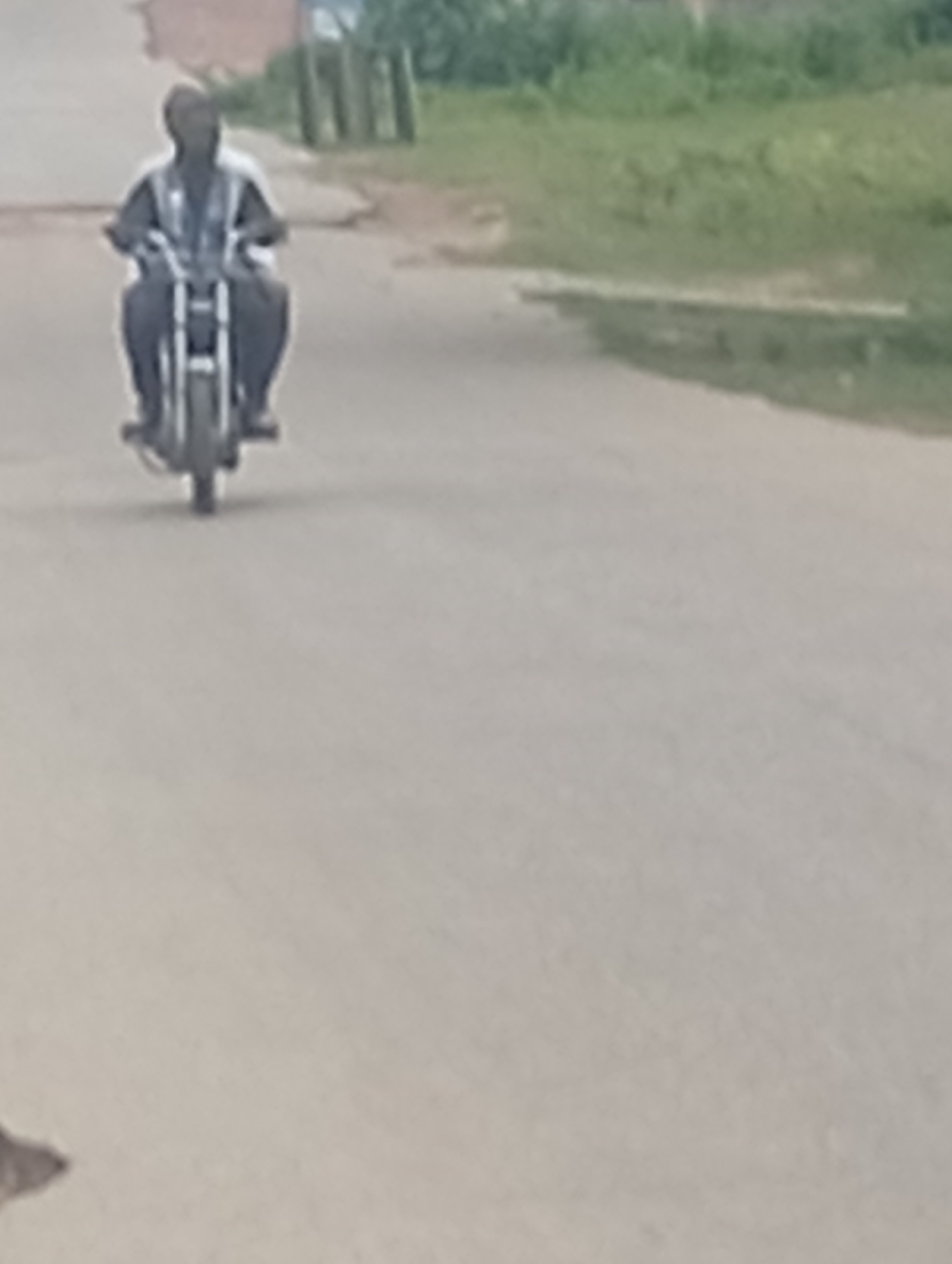 A person is riding a motorcycle down a paved road in Adankolo, Nigeria. The person is wearing a white shirt and dark pants. The motorcycle appears to be black and white. The road is asphalt and there is a grassy area on the right side of the image. There are some buildings in the background. The image is slightly blurry.