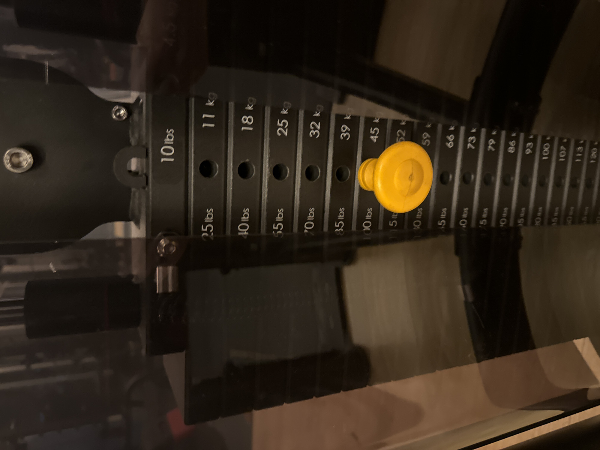 This media file shows a close-up, slightly tilted, vertical view of a weight stack on a home gym machine. The dominant object is the black weight stack, featuring a series of flat, rectangular metal plates. Each plate has an identifying label printed on its side, indicating weight in pounds (lbs) and kilograms (kg). Some of the visible weights include "10 lbs", "11 kg", "25 lbs", "40 lbs", "18 kg", "55 lbs", "25 kg", "32 kg", "39 kg", "45 kg", "52 kg", "59 kg", "66 kg", "73 kg", "79 kg", "86 kg", "93 kg", "100 kg", "107 kg", "113 kg", and "120 kg". A bright yellow pin, likely used to select the desired weight, is inserted into the stack. The background is dark and out of focus, suggesting an indoor setting, possibly a home gym. There are reflections visible on a shiny surface in the foreground, indicating a protective enclosure or glass panel. The lighting appears to be artificial and somewhat dim, casting shadows. No people or specific location cues related to Murcia, Spain are discernible in the image. The scene is static and focuses solely on the gym equipment.