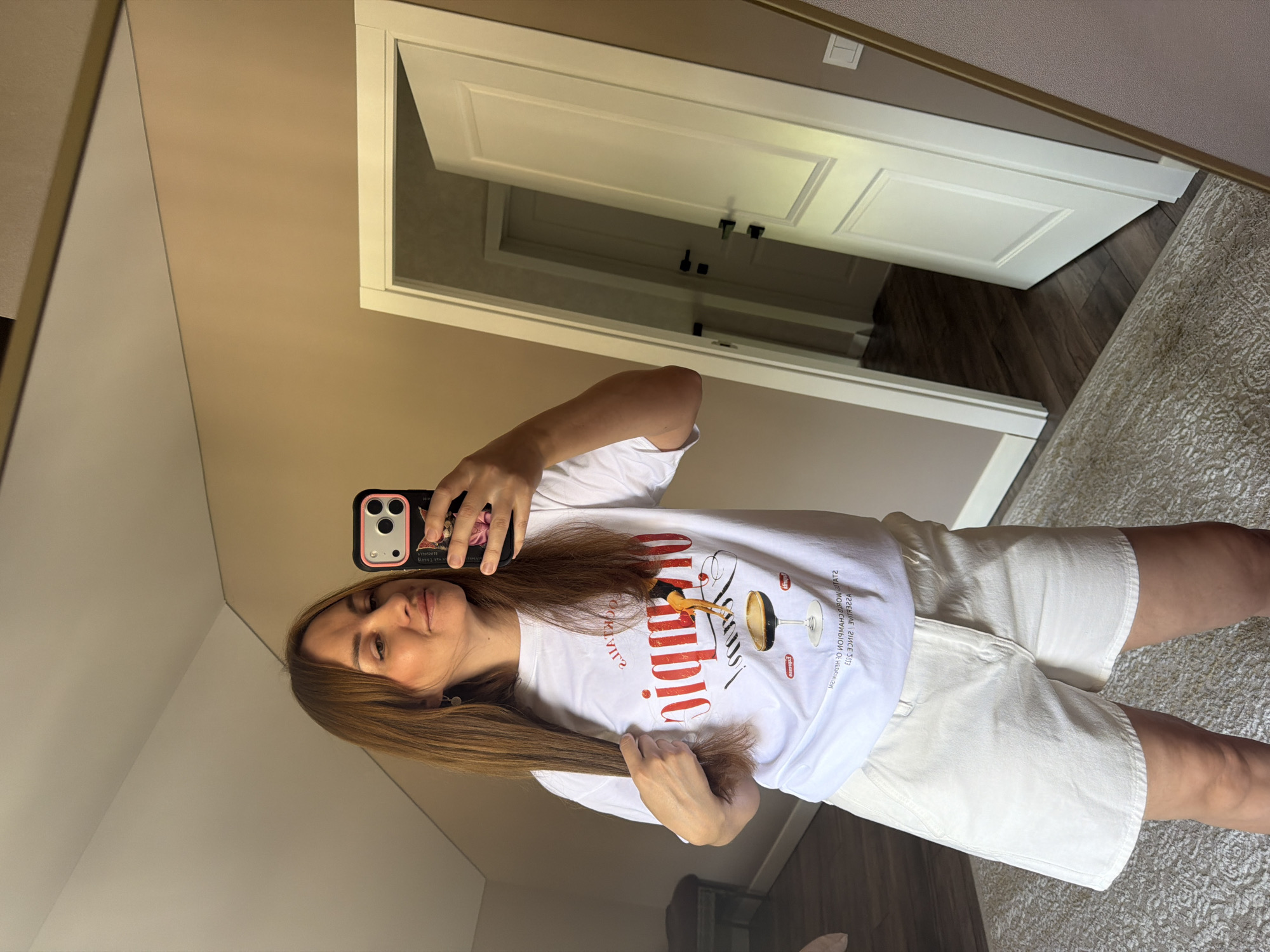 A woman with long, light brown hair is standing in a room, taking a selfie in a mirror. She is wearing a white t-shirt with a graphic print and white denim shorts. Her left hand is holding a section of her hair. In the reflection, a door is visible, slightly ajar, revealing another doorway and part of a hallway. The room appears to be a domestic setting, possibly an apartment or house. The background wall is painted a neutral, light brown color. The floor is covered with a patterned rug and wooden flooring. There are no visible indicators that the location is in Malinovka, Russia.